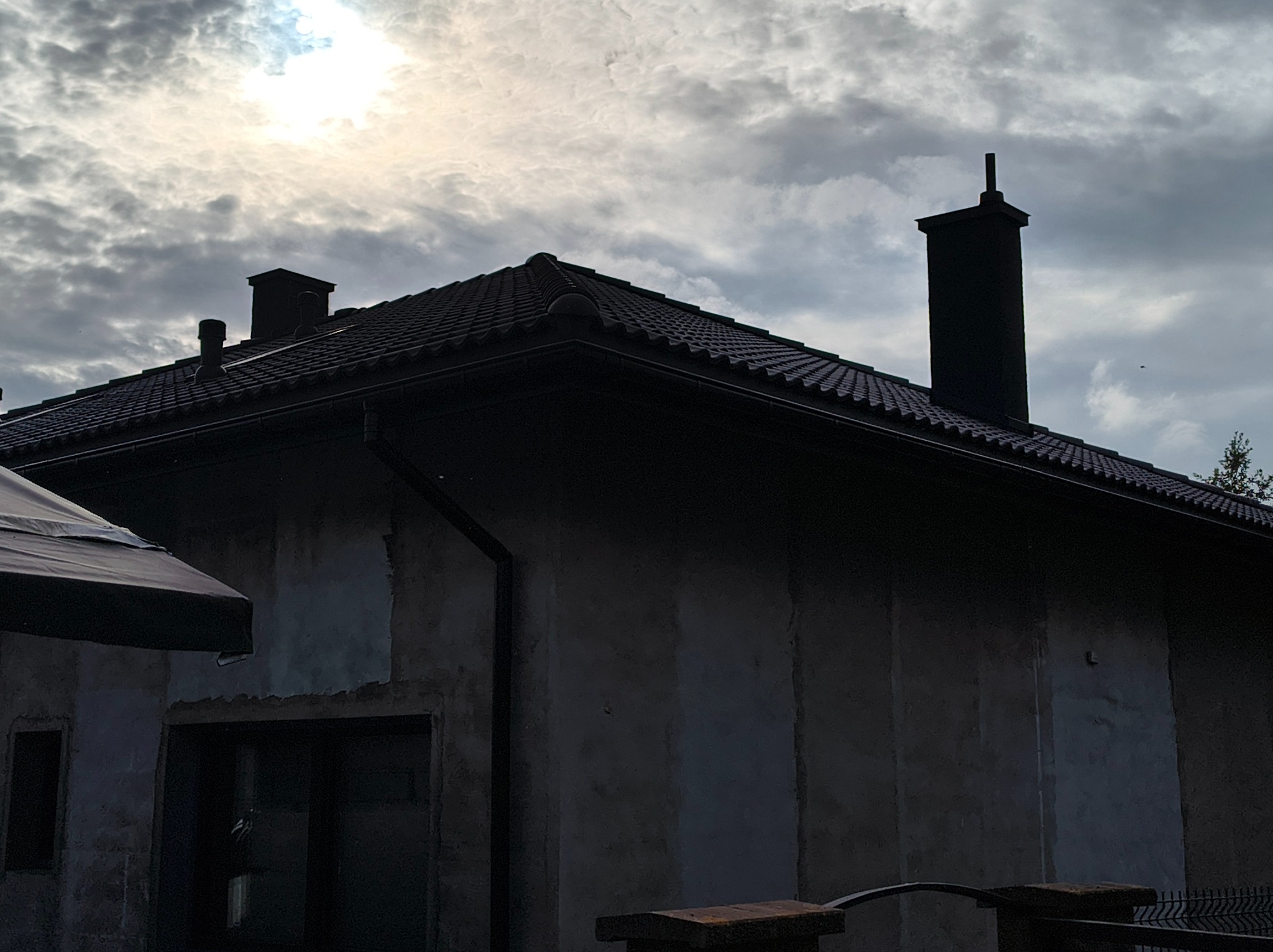 Dom z ciemną dachówką ceramiczną i kominami na tle pochmurnego nieba. Widoczna elewacja w trakcie prac wykończeniowych.