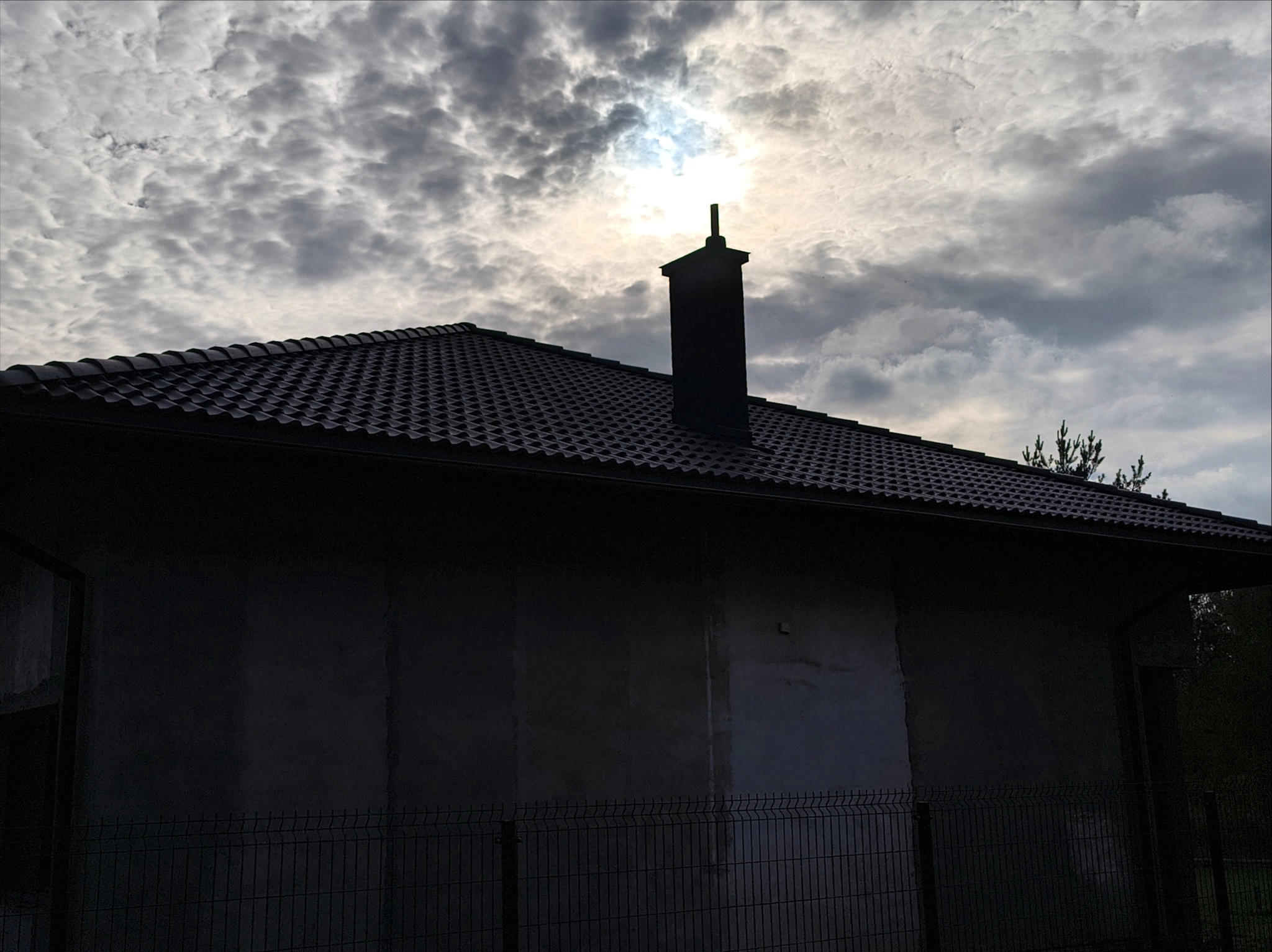 Ciemny dach z kominem na tle pochmurnego nieba. Widok z dołu na fragment budynku w trakcie budowy, ogrodzony siatką. Zastosowano ciemną blachodachówkę.