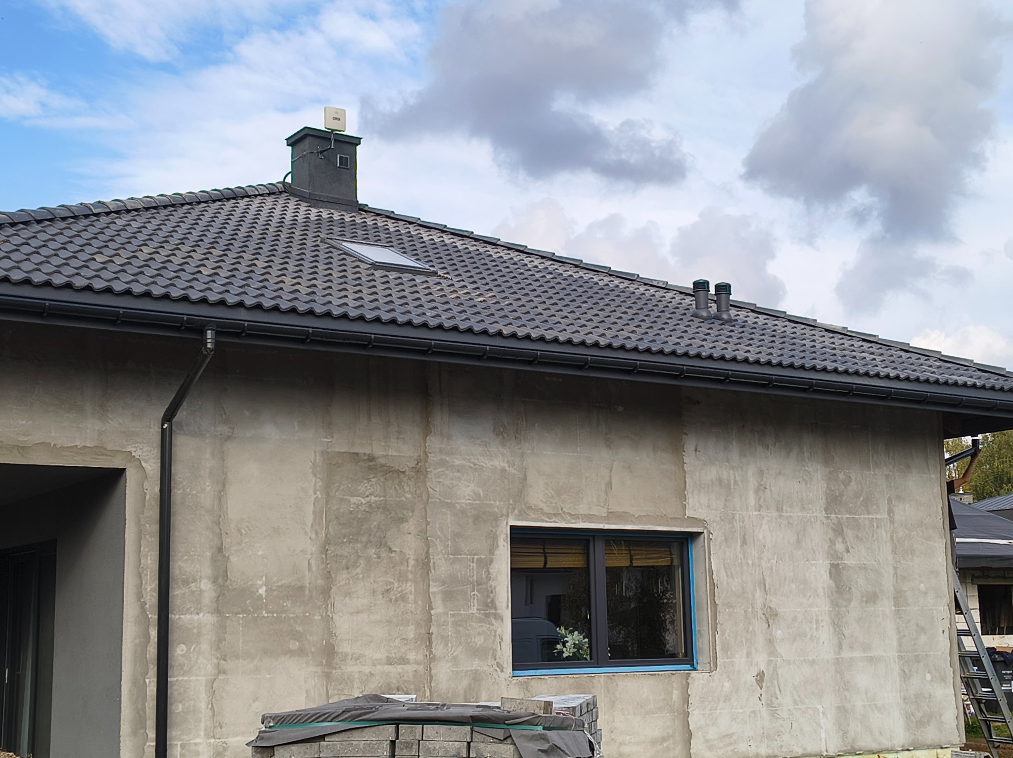 Dom w trakcie prac wykończeniowych: szara dachówka, ciemne rynny, surowa elewacja z oknem. Widoczny komin z anteną i materiały budowlane.