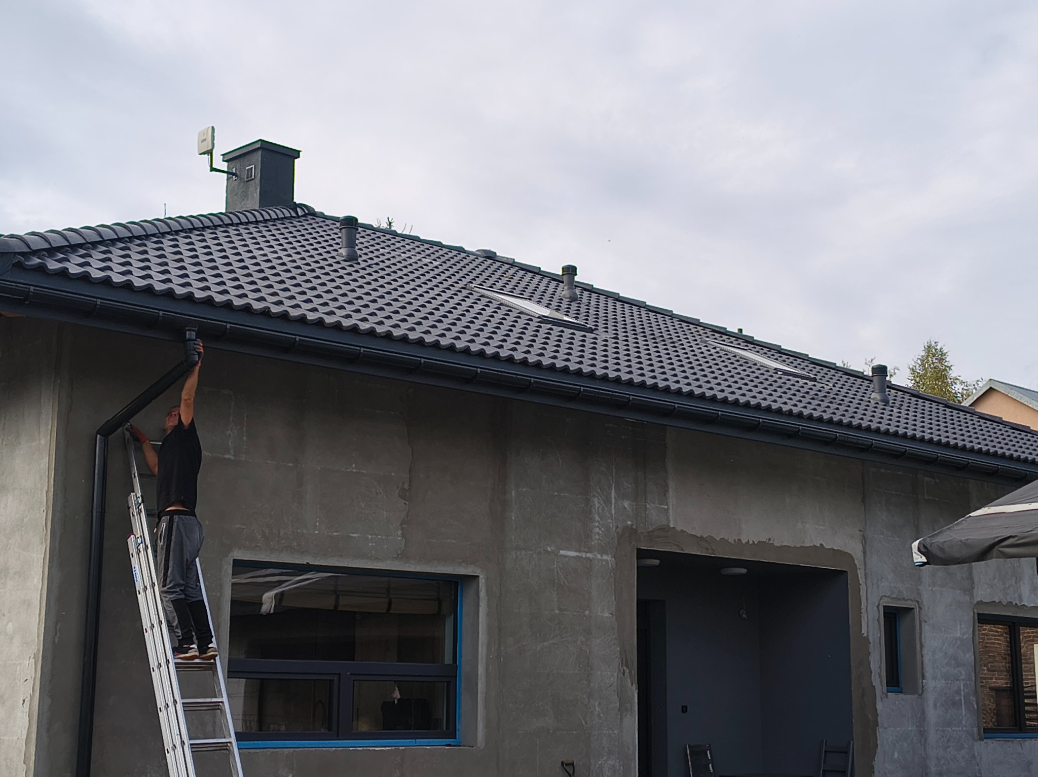 Monter na drabinie instaluje rynnę na tle surowej elewacji domu z ciemnoszarym dachem. Widoczne okna z niebieską taśmą zabezpieczającą.