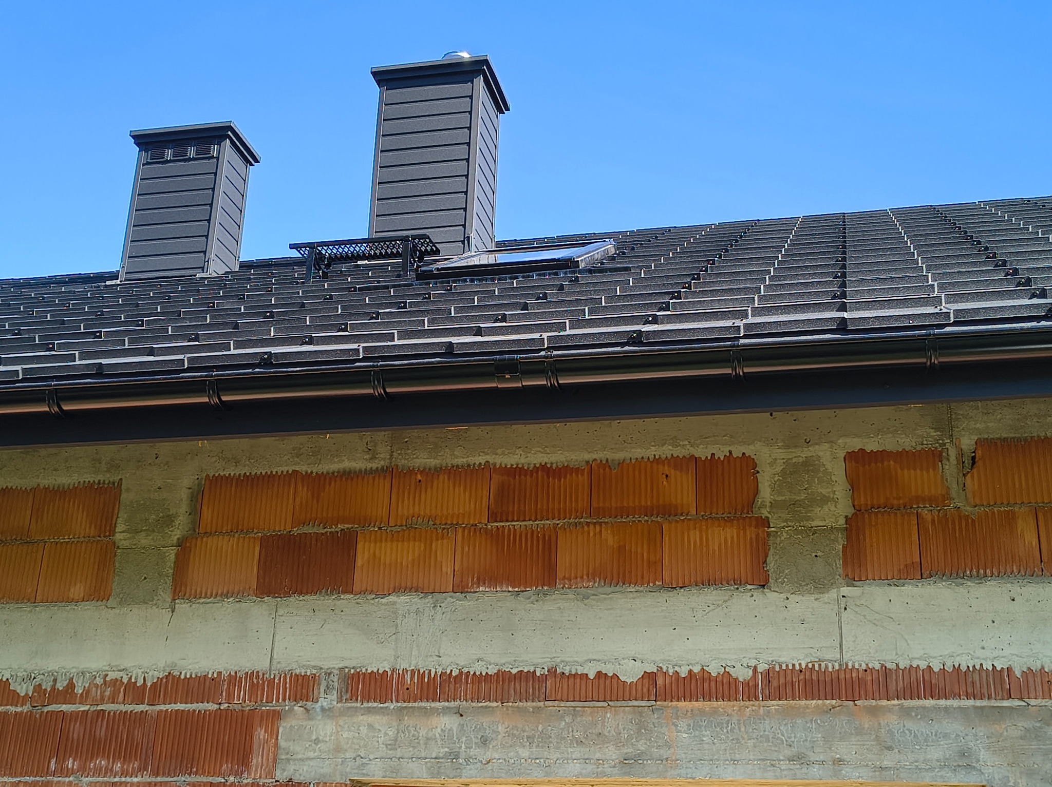 Ciemnoszary dach z dwoma kominami i oknem dachowym, widoczny fragment ściany z czerwonej cegły i szarego betonu w słoneczny dzień.