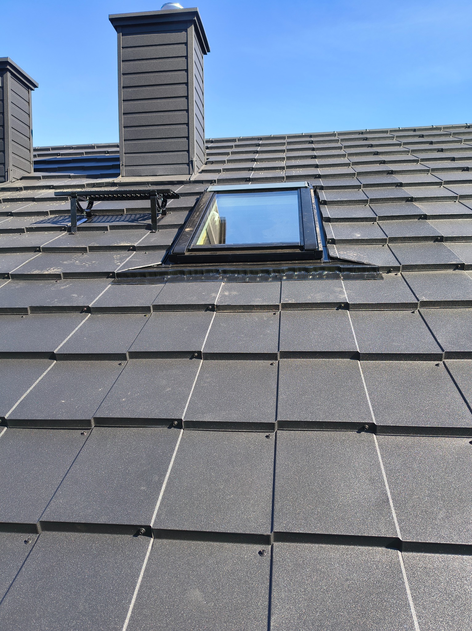 Grafitowy dach z oknem połaciowym i kominami w słoneczny dzień. Widoczny fragment dachu z drabinką kominiarską. Nowoczesne pokrycie dachowe.