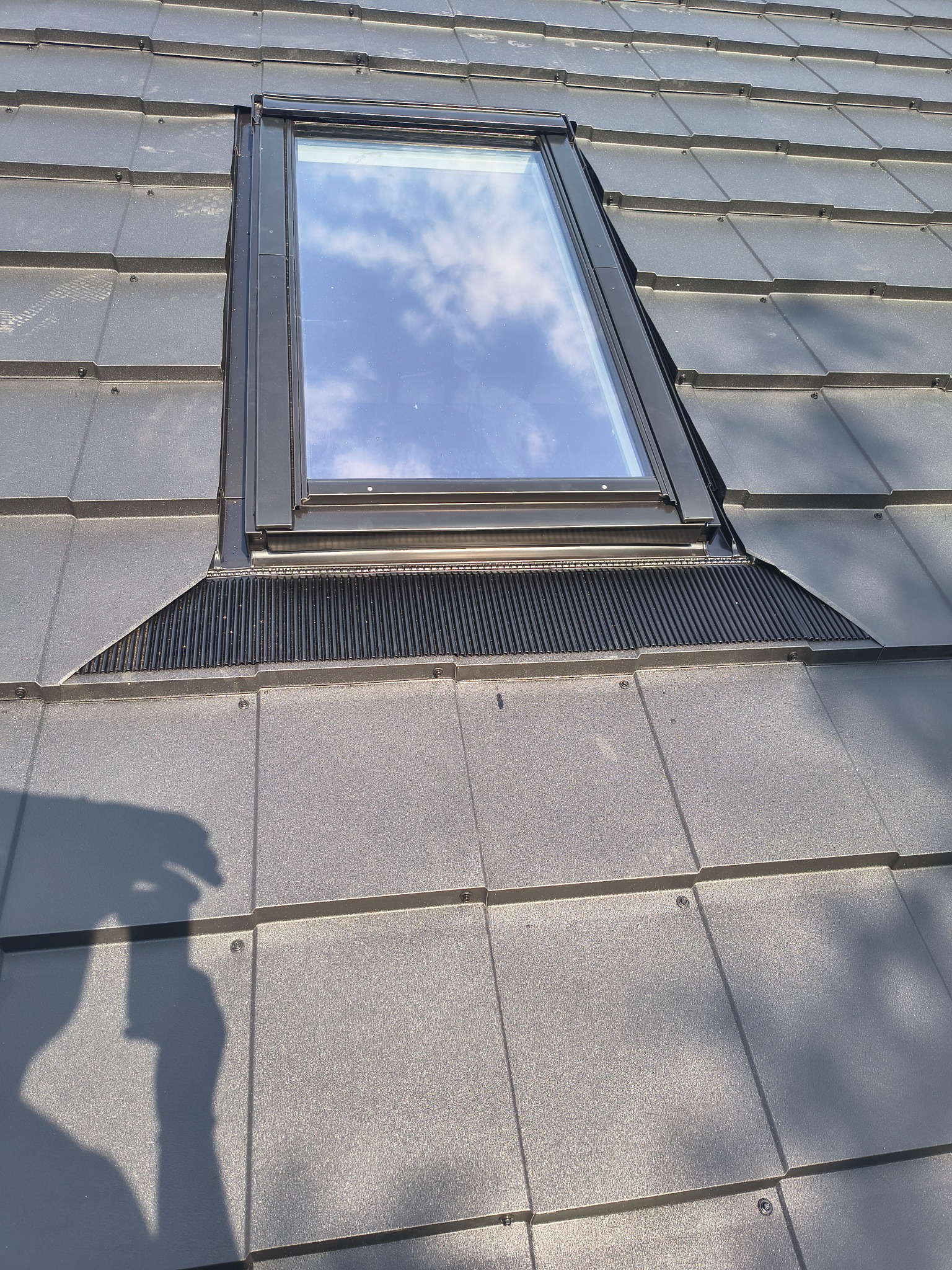 Szara dachówka ceramiczna z zamontowanym oknem dachowym, w którym odbijają się chmury. Widoczny element wentylacyjny pod oknem i cień osoby robiącej zdjęcie.