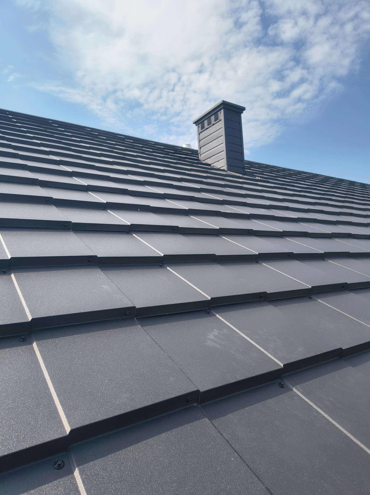 Szary, metalowy dach z kominem na tle błękitnego nieba z chmurami. Widok z dołu ukazuje regularne ułożenie dachówek i estetyczne wykończenie komina.