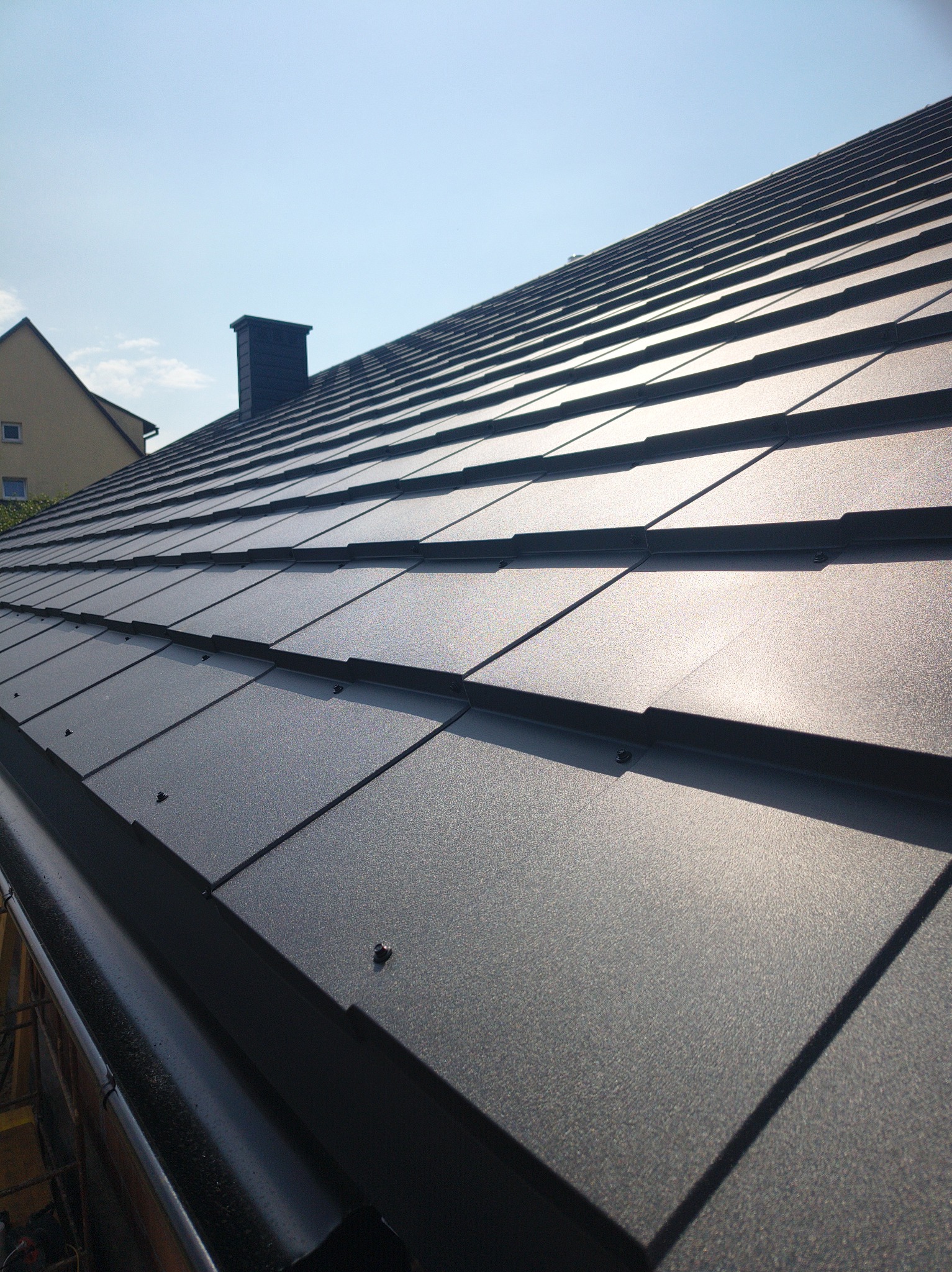 Nowy, grafitowy dach z blachodachówki modułowej w promieniach słońca, z widoczną rynną i kominem w tle. Detal łączenia arkuszy, minimalizm.