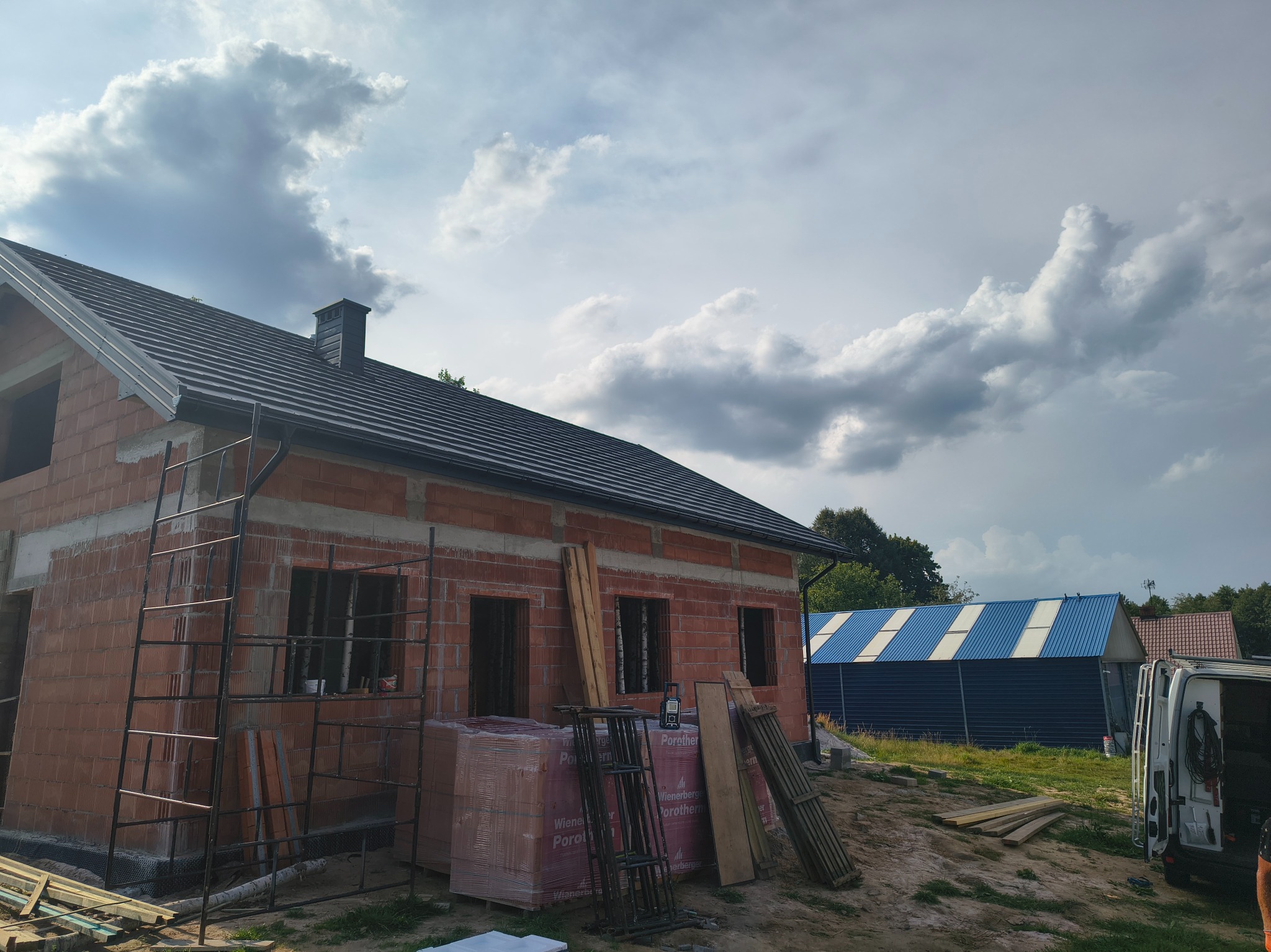 Surowy budynek z cegły Porotherm z ciemnym dachem, widoczne rusztowanie, palety z cegłami i częściowo w tle budynek z blachy trapezowej.