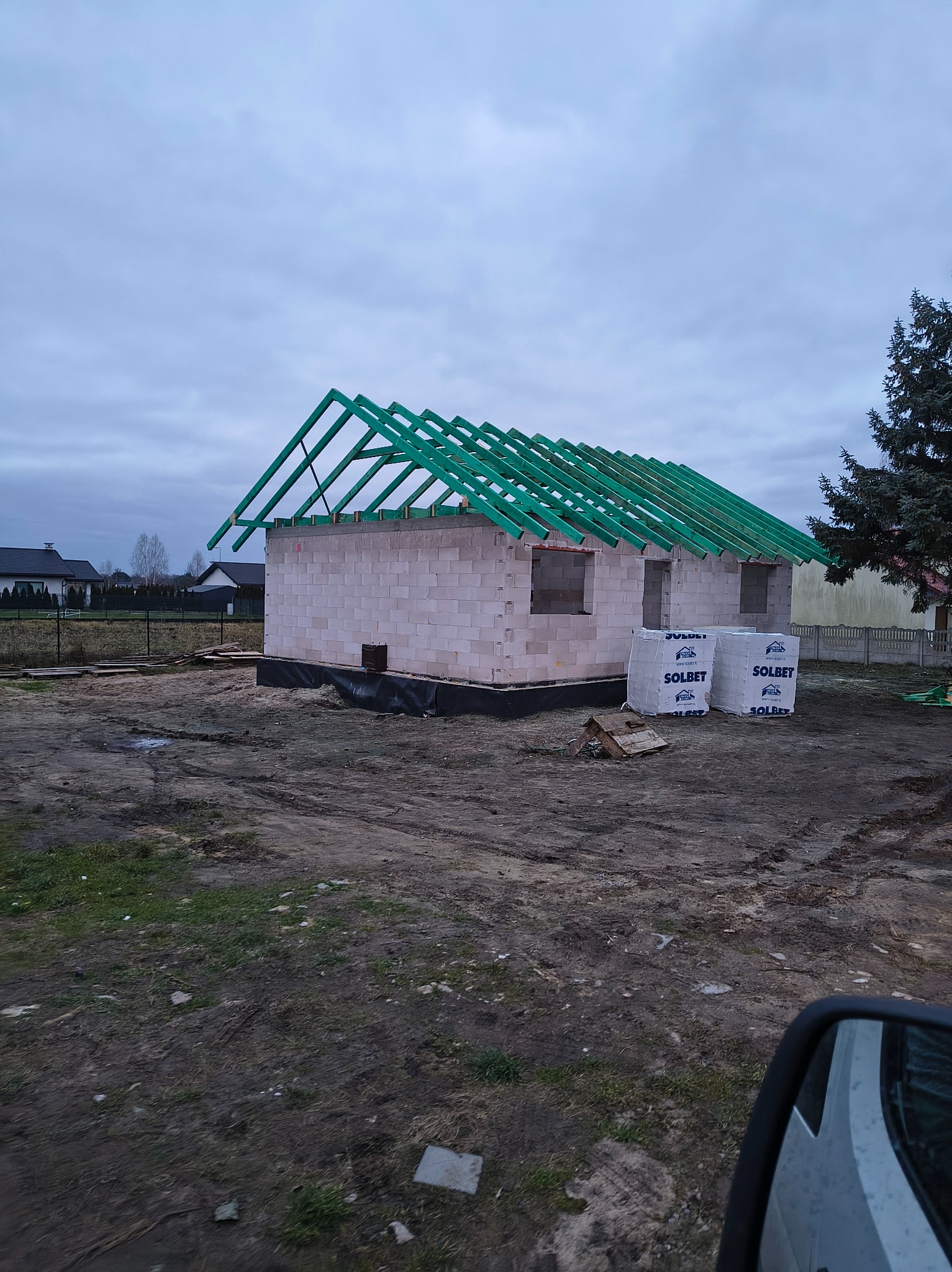 Budowa domu jednorodzinnego w stanie surowym otwartym z zieloną konstrukcją dachu i paletami bloczków Solbet na działce. Pochmurne niebo w tle.