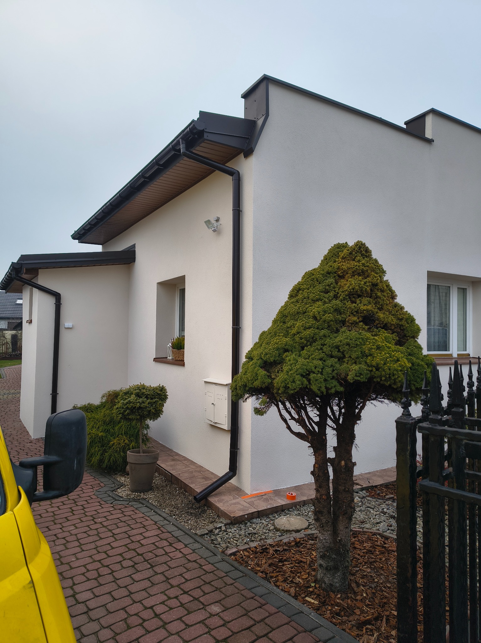 Elewacja domu jednorodzinnego po ociepleniu, z widocznymi rynnami w kolorze grafitowym. Uwagę zwraca równa, jasna powierzchnia ściany i zadbany ogródek.