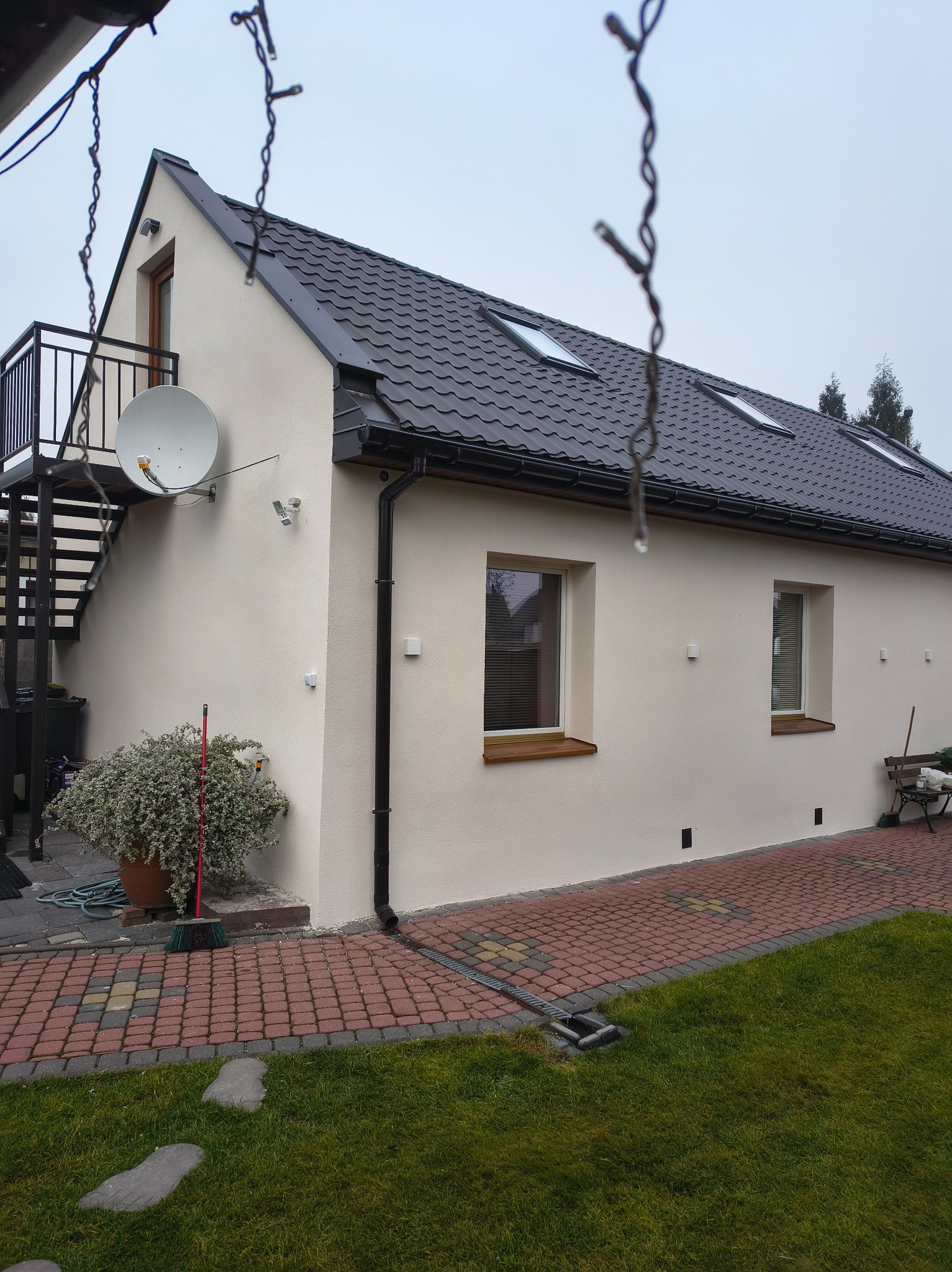 Elewacja budynku z ciemną dachówką, rynnami i schodami zewnętrznymi. Widoczne okna z drewnianymi parapetami, trawnik i ścieżka z kostki.