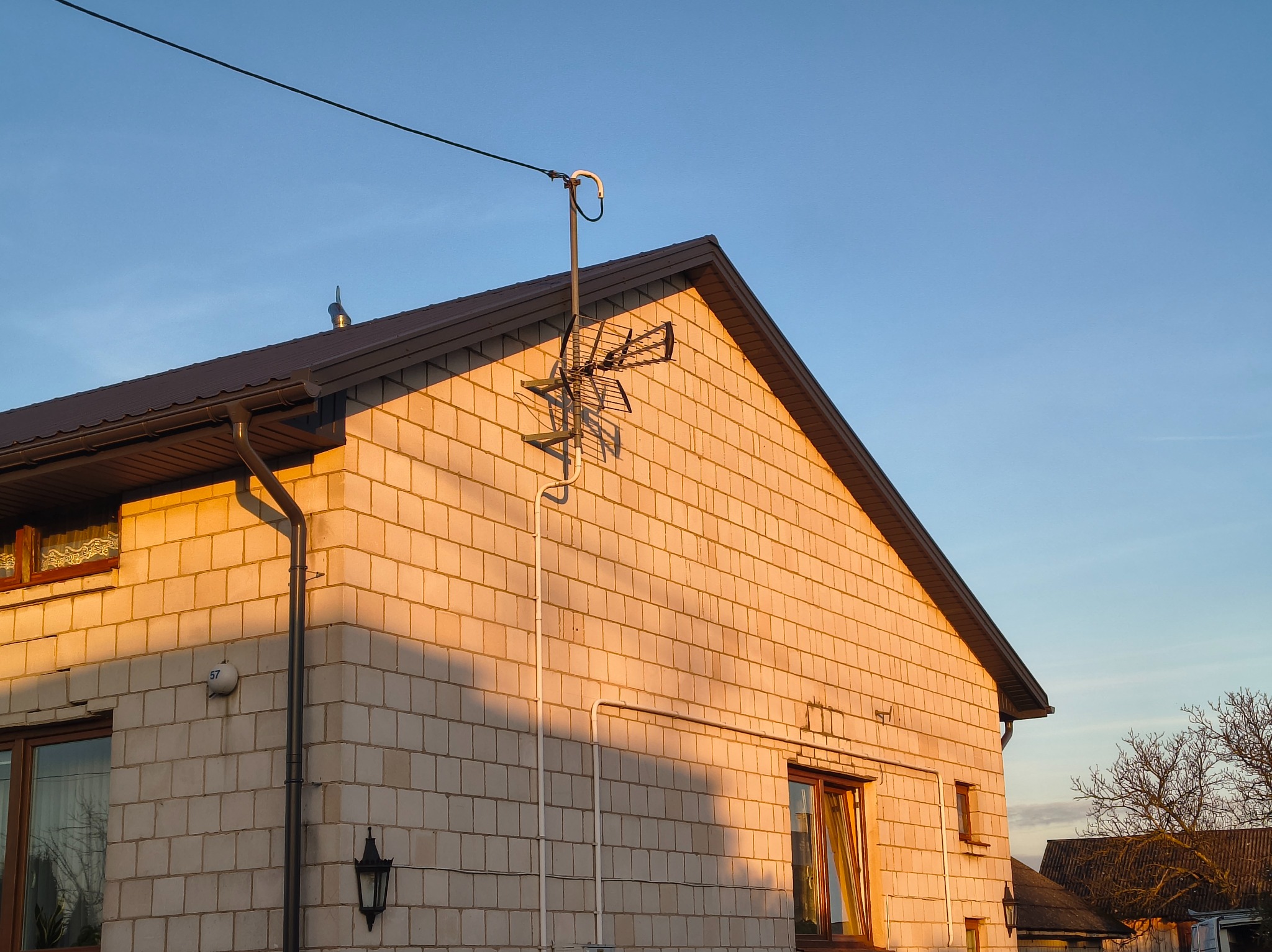 Dwuspadowy dach z brązową blachodachówką i rynnami, oświetlony ciepłym światłem zachodzącego słońca. Na ścianie anteny, widoczne okna i lampa zewnętrzna.