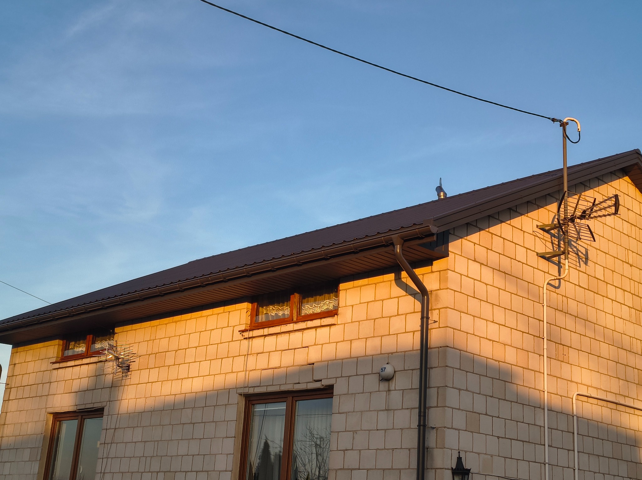 Dom z brązowym dachem i elewacją z jasnej cegły w ciepłym świetle zachodzącego słońca. Widoczne rynny, okna z drewnianymi ramami i antena na dachu.