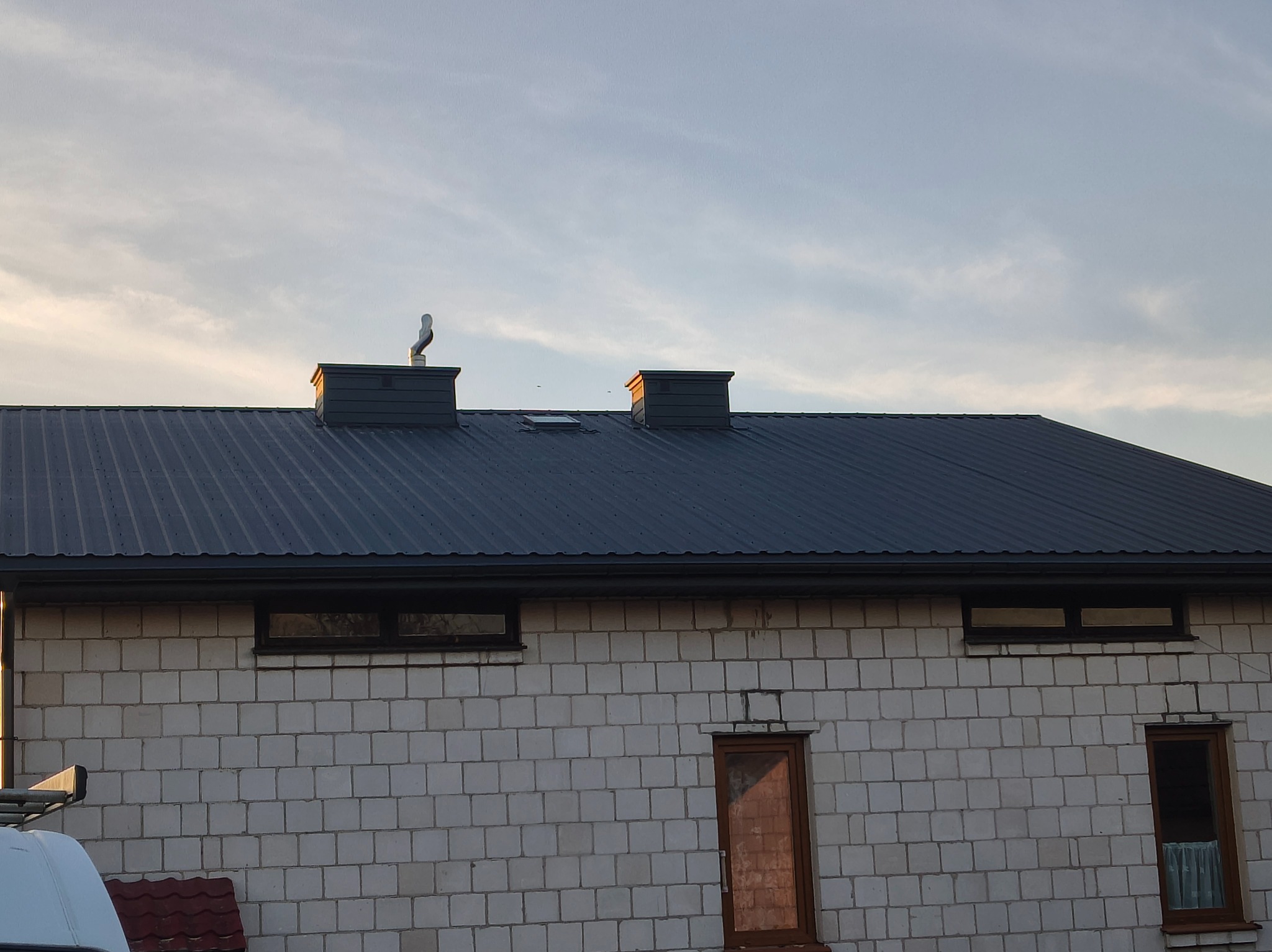 Ciemnoszary dach z blachy trapezowej na budynku z białego pustaka, widoczne kominy i okno dachowe. Ujęcie z dołu, niebo w tle.