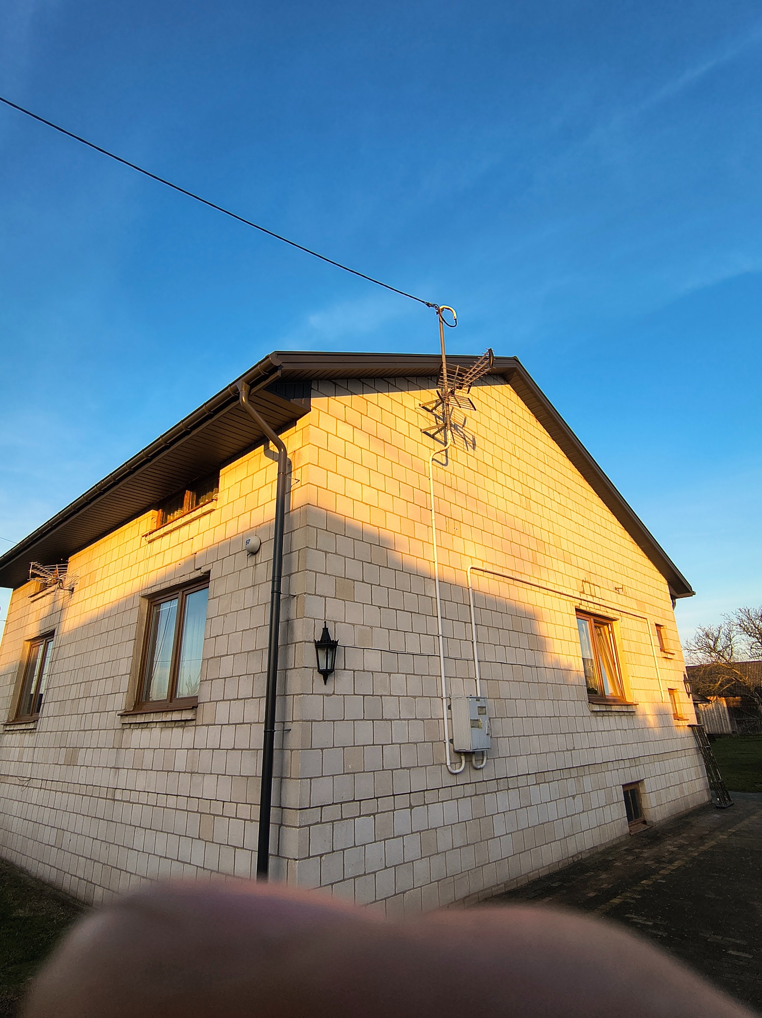 Dom z jasnej cegły w promieniach zachodzącego słońca, widok z dołu. Widoczne okna, rynny, antena na dachu i fragment instalacji elektrycznej na elewacji.