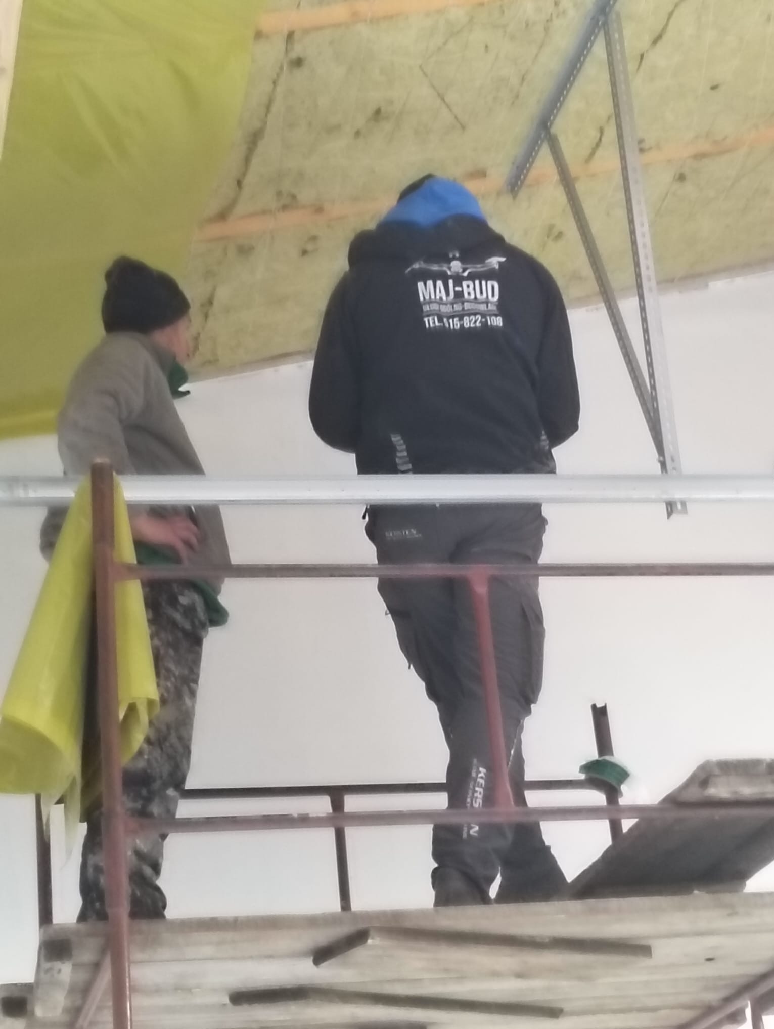 Pracownicy na rusztowaniu montują ocieplenie poddasza, widoczna wełna mineralna i folia paroizolacyjna. Jeden z pracowników w bluzie z logo firmy.