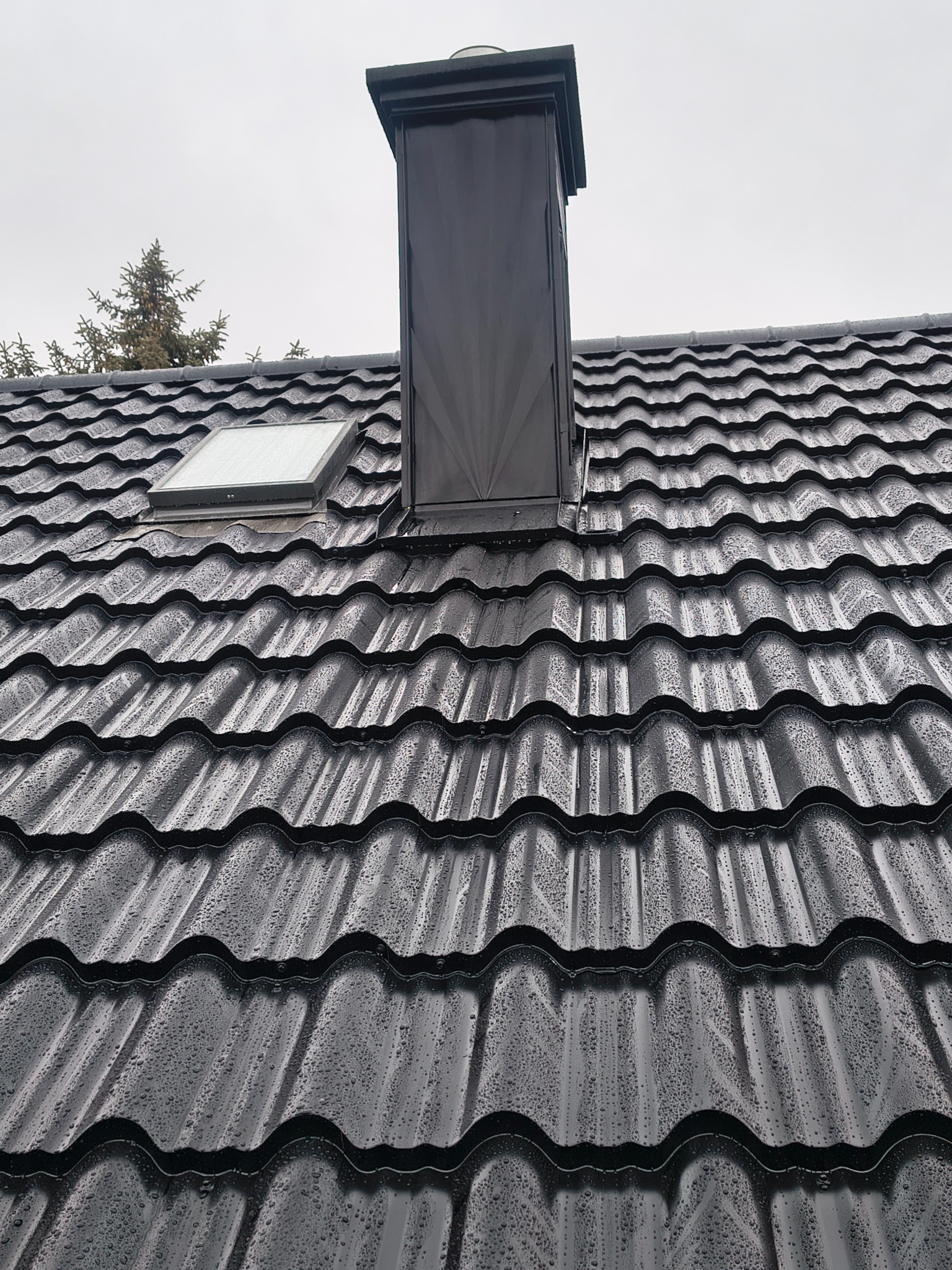 Czarny dach z blachodachówki modułowej, pokryty kroplami deszczu, z oknem dachowym i kominem. Widok z dołu na szarym tle nieba.