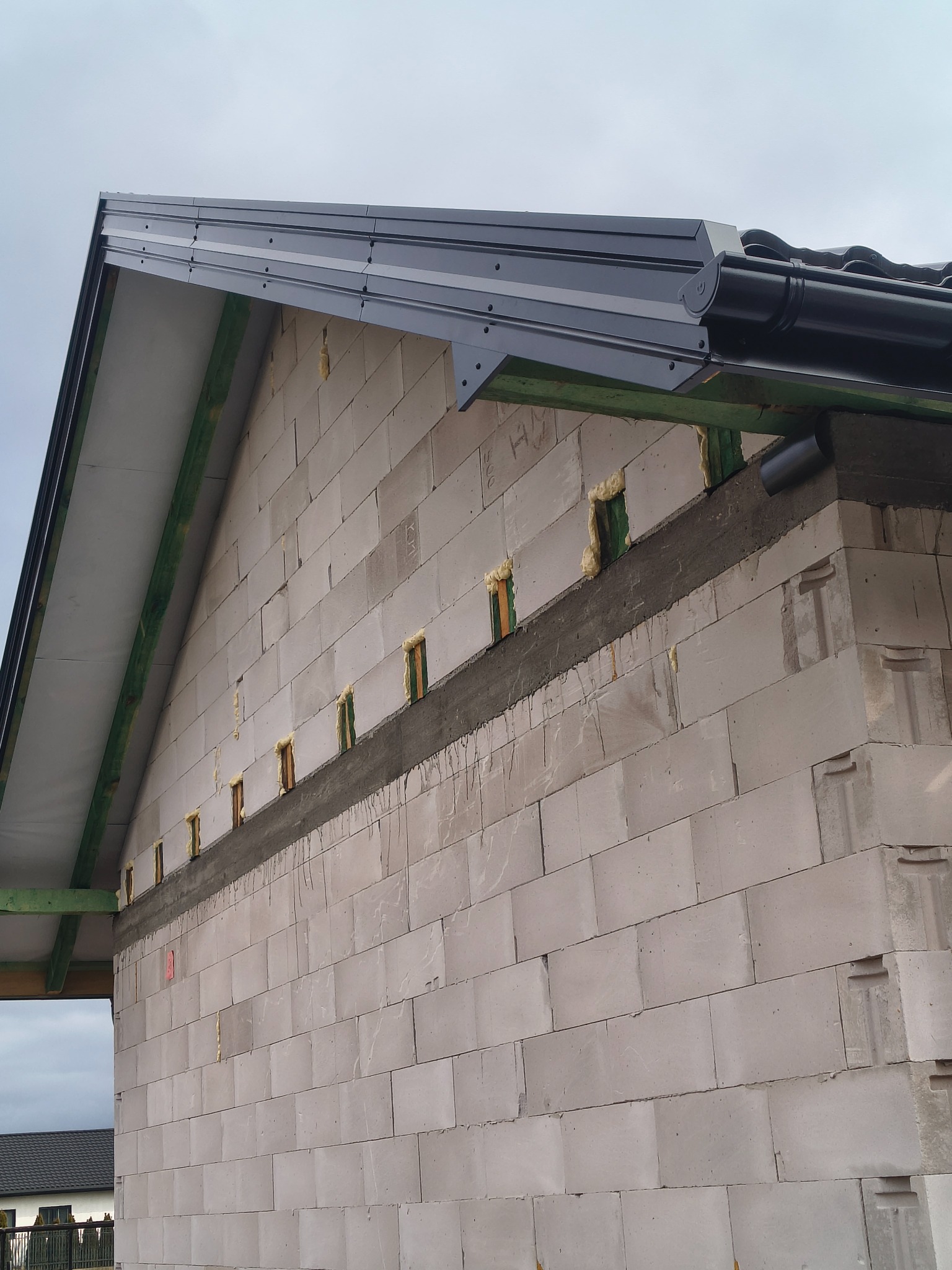 Szara blacha na dachu budynku z betonu komórkowego. Widoczne rynny i pianka montażowa w otworach. Elementy drewniane konstrukcji dachu w kolorze zielonym.