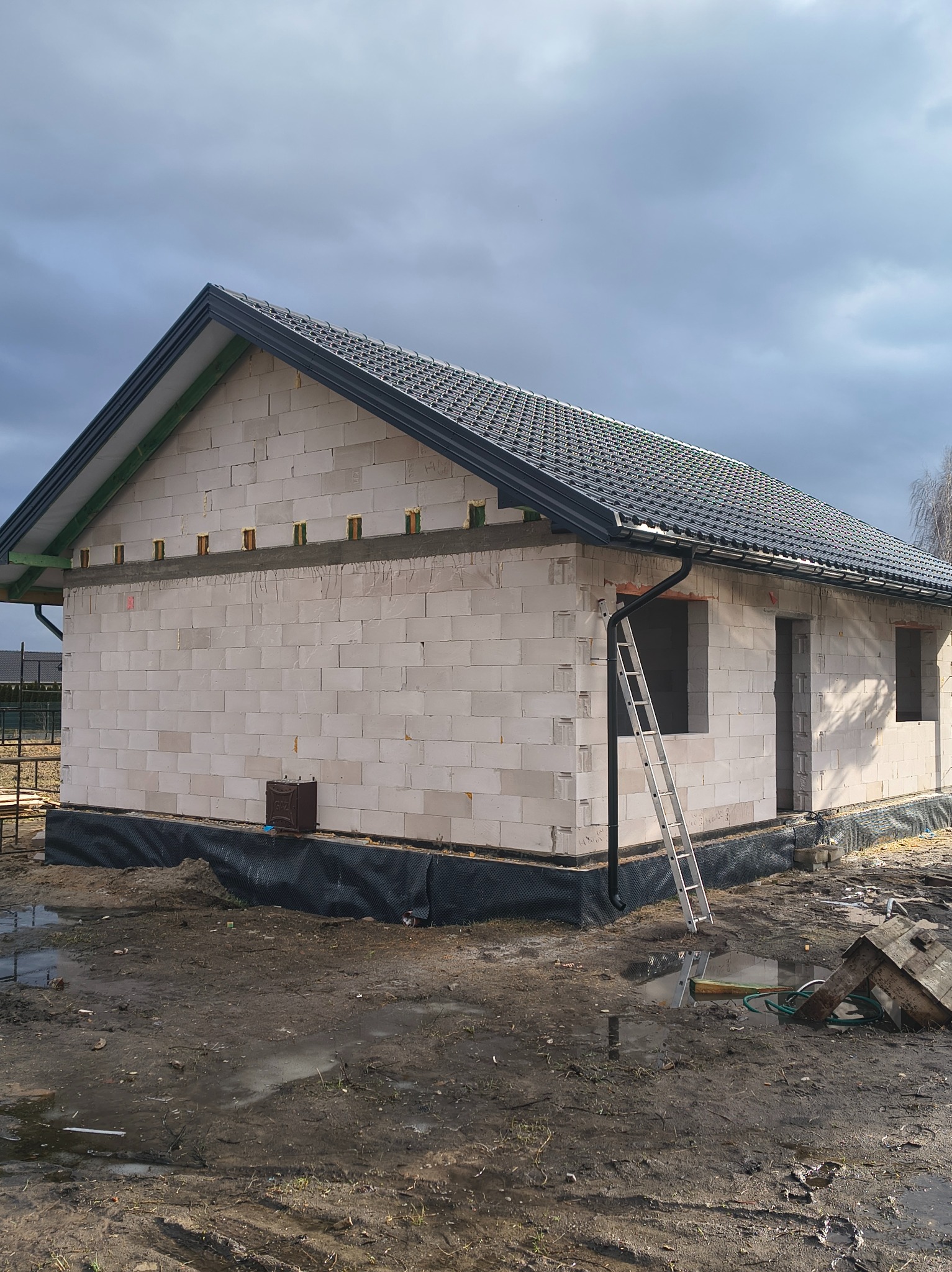 Niedokończony dom z białego bloczka z czarnym dachem z dachówki, drabina oparta o rynnę, widoczne otwory okienne i drzwiowe, teren wokół domu błotnisty.