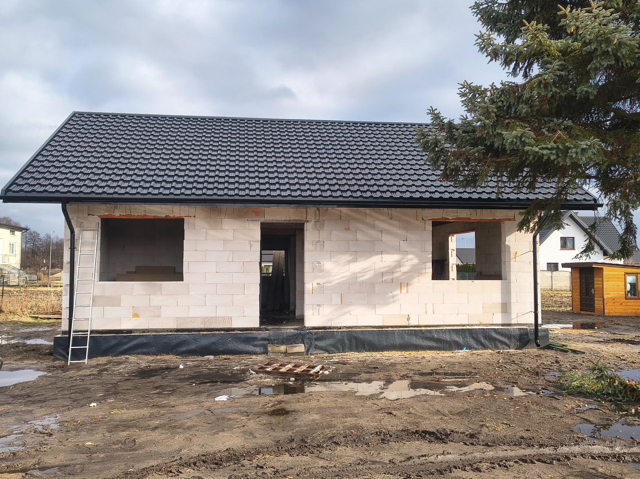 Surowy stan domu jednorodzinnego z czarnym dachem z blachodachówki, widoczne otwory okienne i drzwiowe, drabina oparta o ścianę, teren wokół budynku błotnisty.