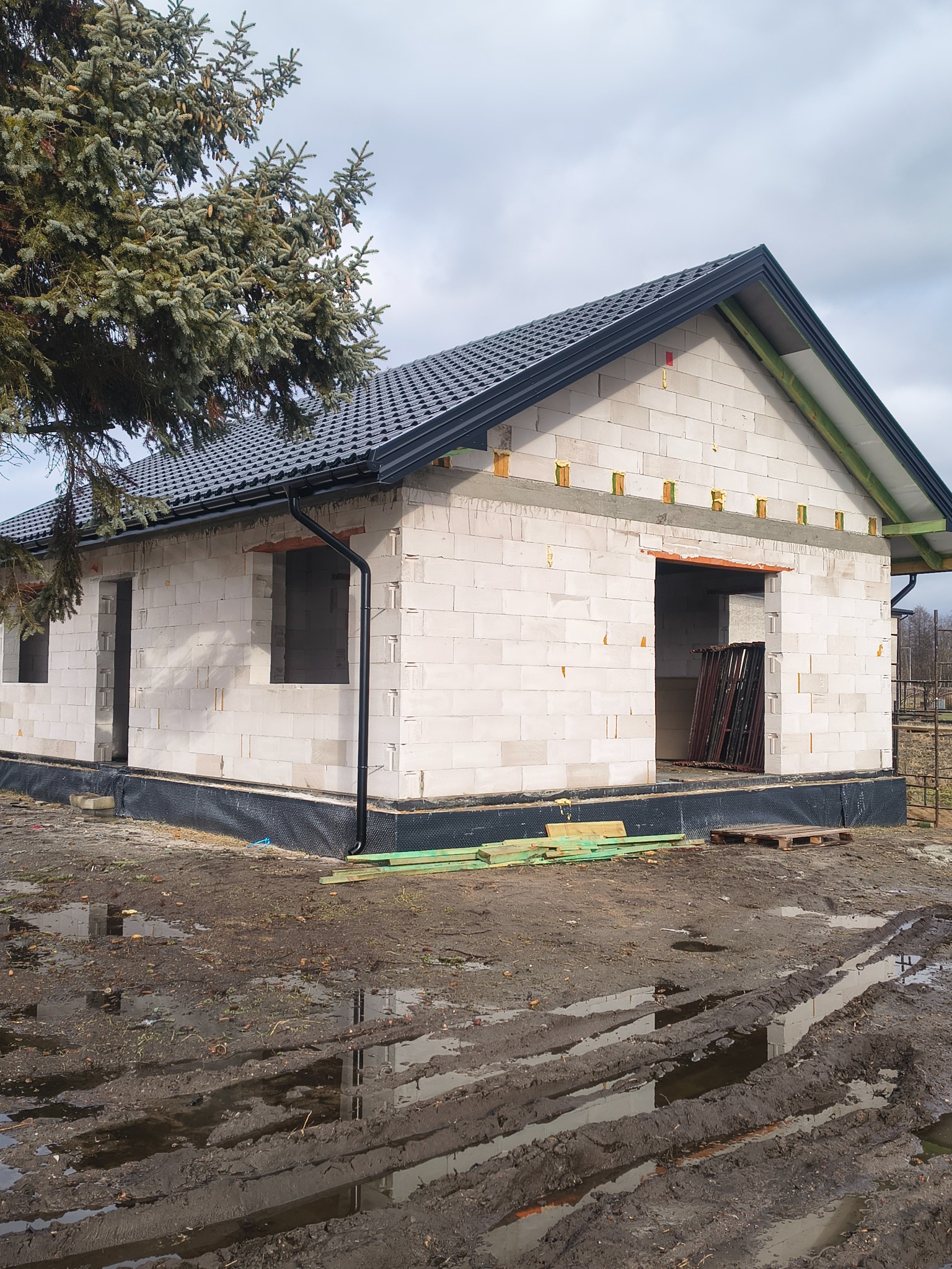 Surowy stan domu z bloczków, z czarnym dachem i rynnami, widoczne okna i drzwi w stanie surowym, teren błotnisty, drzewo iglaste z lewej strony.