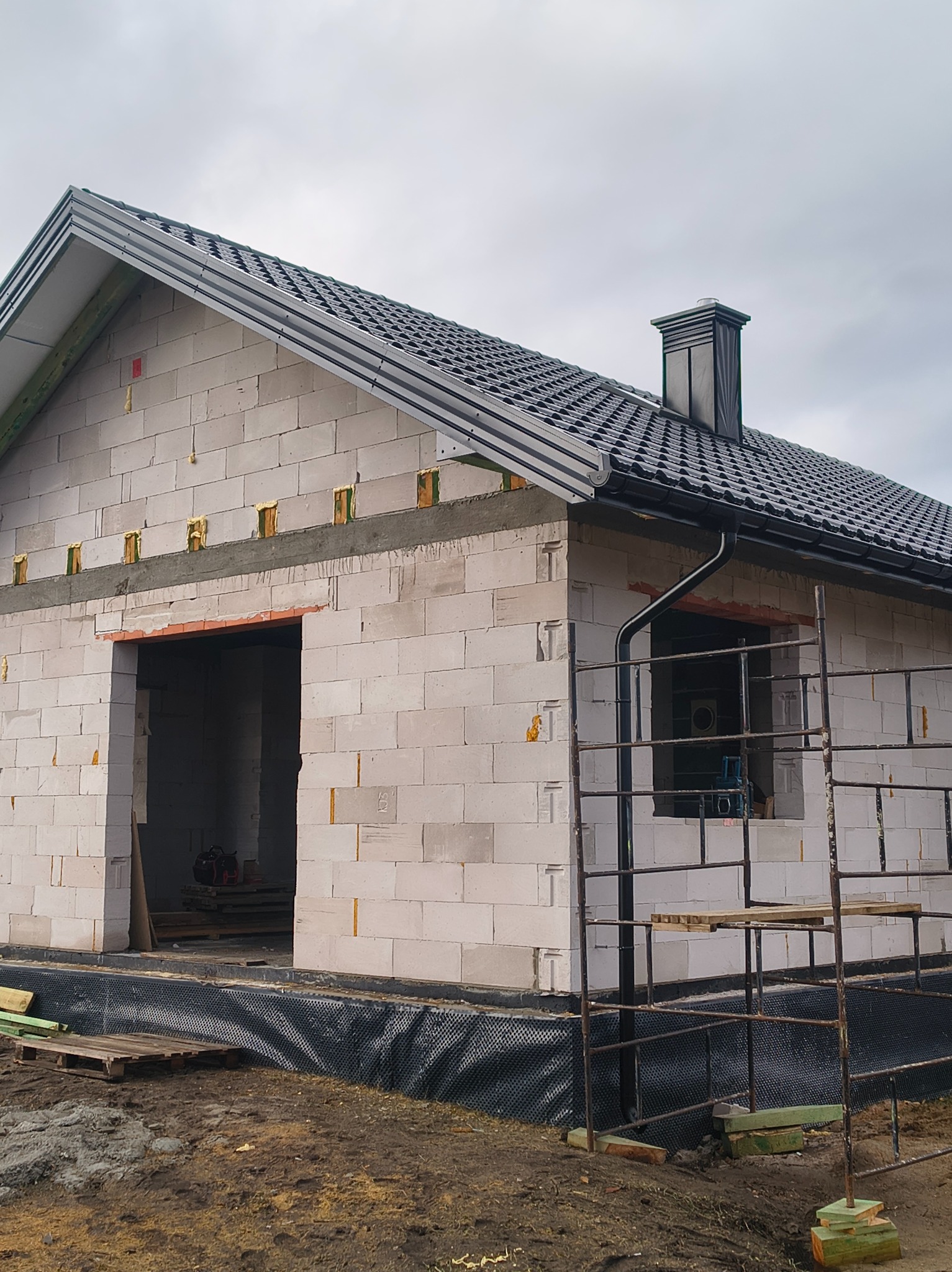 Dom w trakcie budowy z czarną dachówką i rynnami. Widoczne białe bloczki, rusztowania i elementy izolacji. Pochmurne niebo w tle.