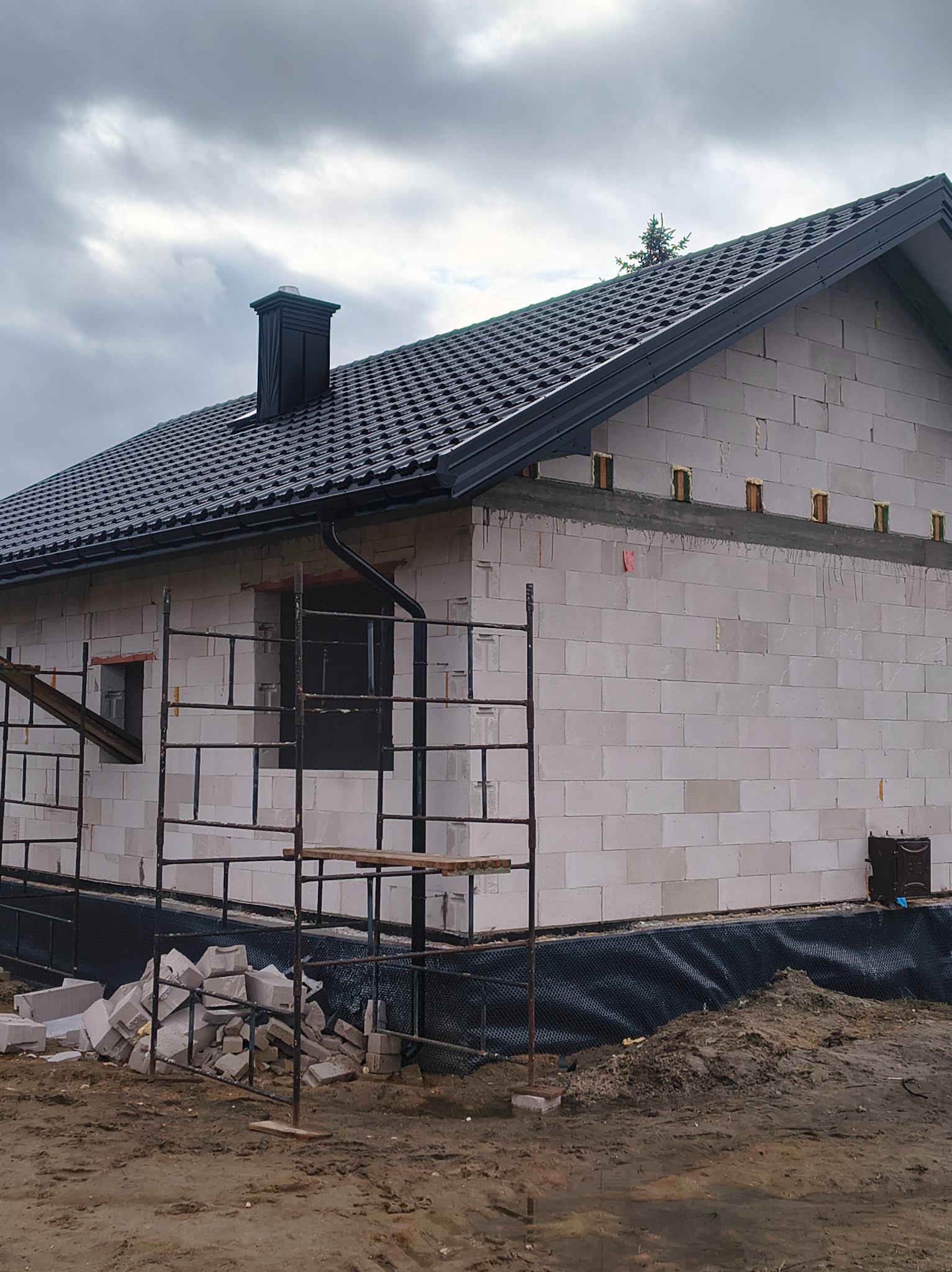 Dom w trakcie budowy z czarnym dachem i kominem, widoczne rusztowania i materiały budowlane. Ujęcie z boku, pochmurne niebo w tle.