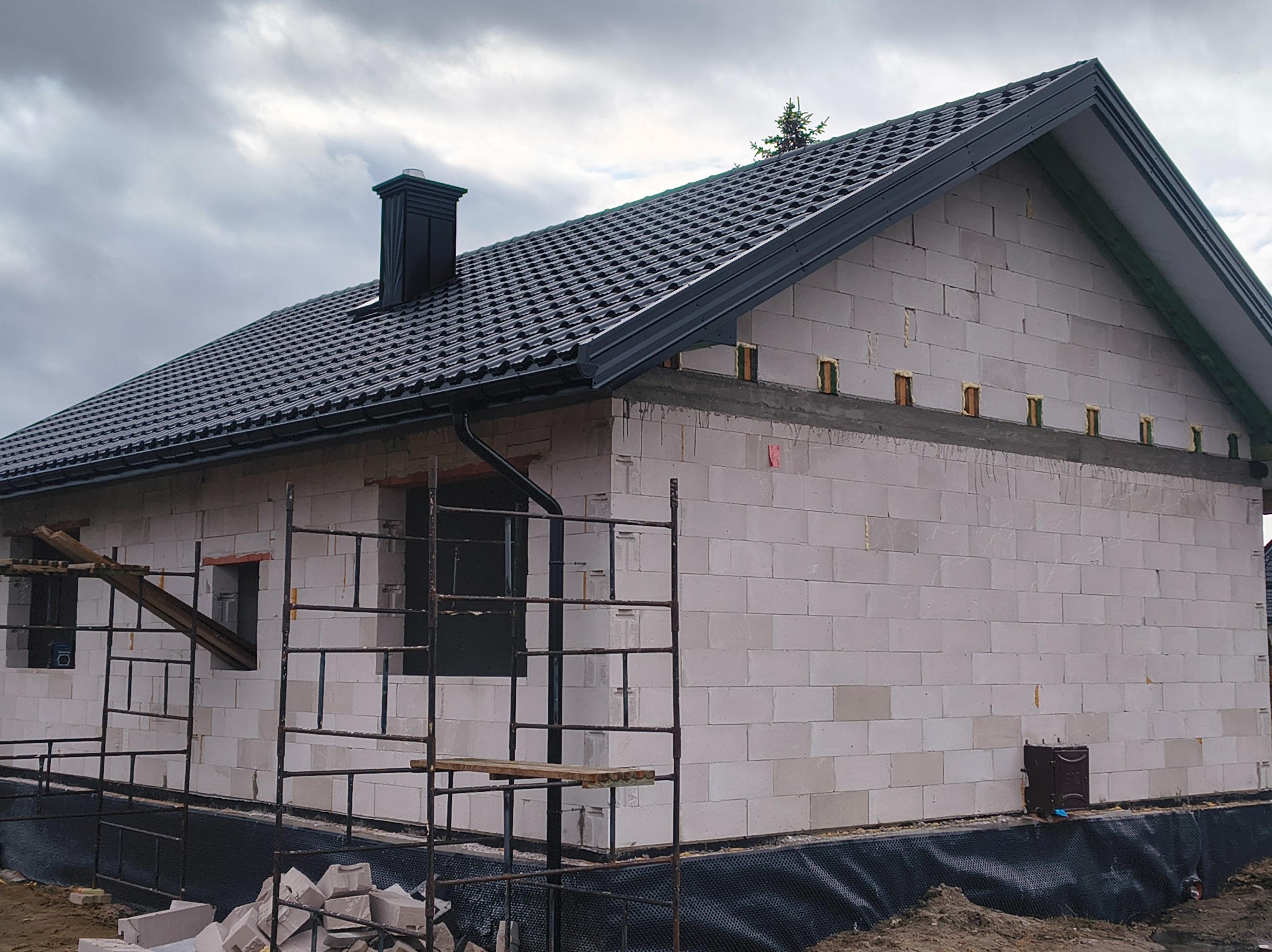 Dom w trakcie budowy z czarnym dachem z blachy trapezowej i kominem. Widoczne rusztowania i bloczki z betonu komórkowego. Pochmurne niebo w tle.