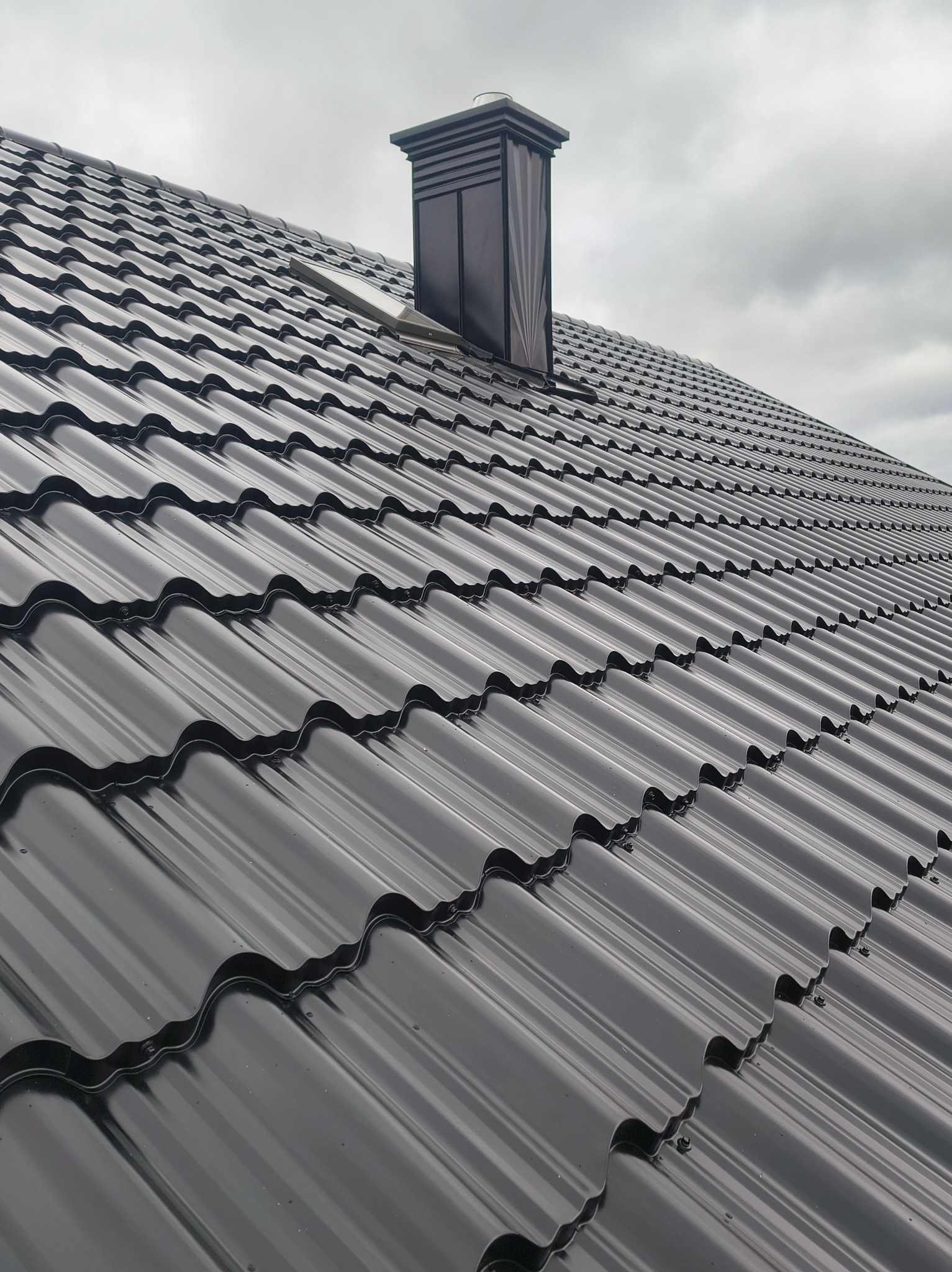 Czarny dach z blachy trapezowej z kominem wentylacyjnym na tle pochmurnego nieba. Widok z dołu, ukazujący regularny wzór pokrycia.