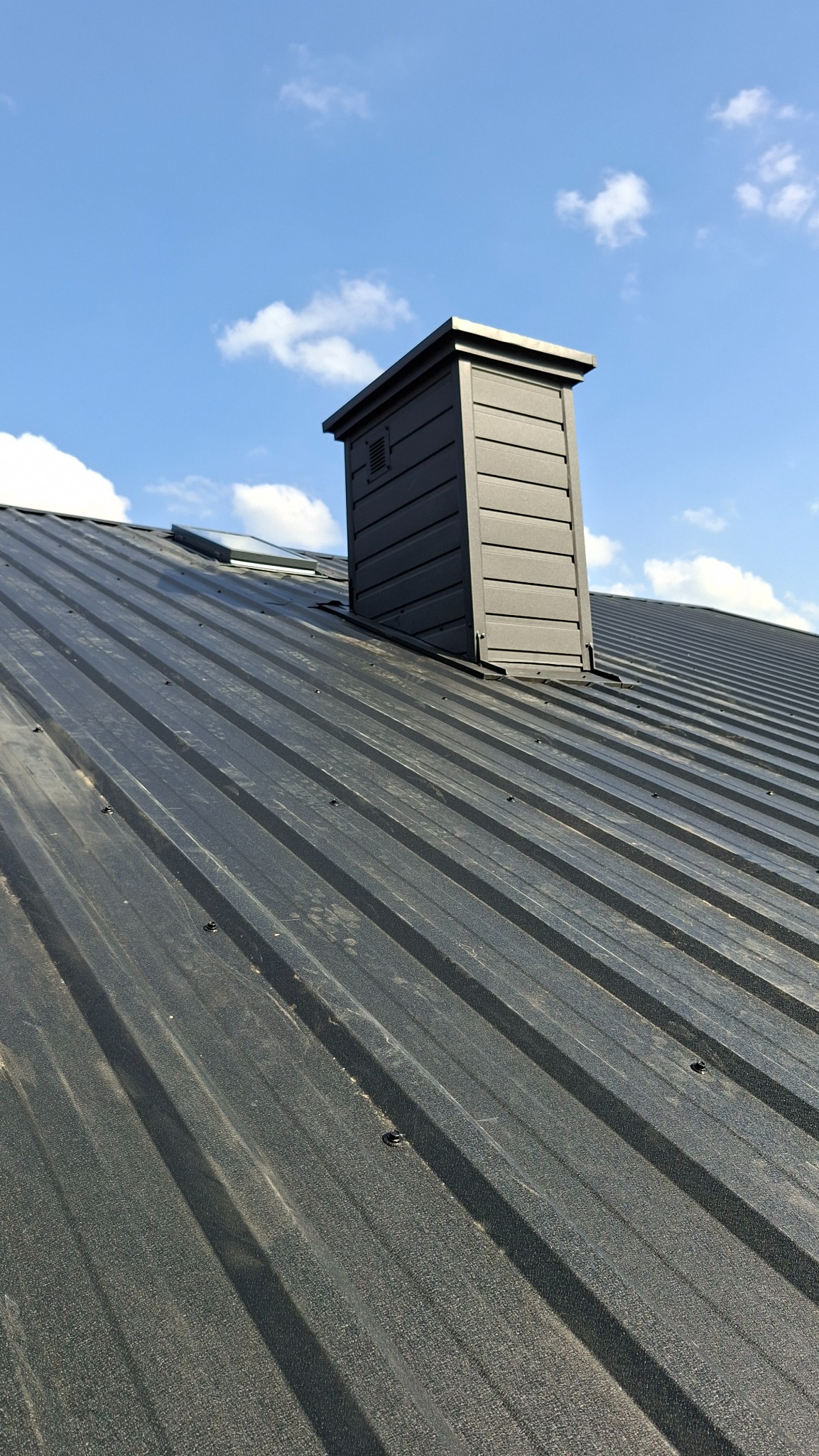 Grafitowy dach z blachy trapezowej z kominem wentylacyjnym i oknem dachowym na tle błękitnego nieba z chmurami. Widok z dołu.
