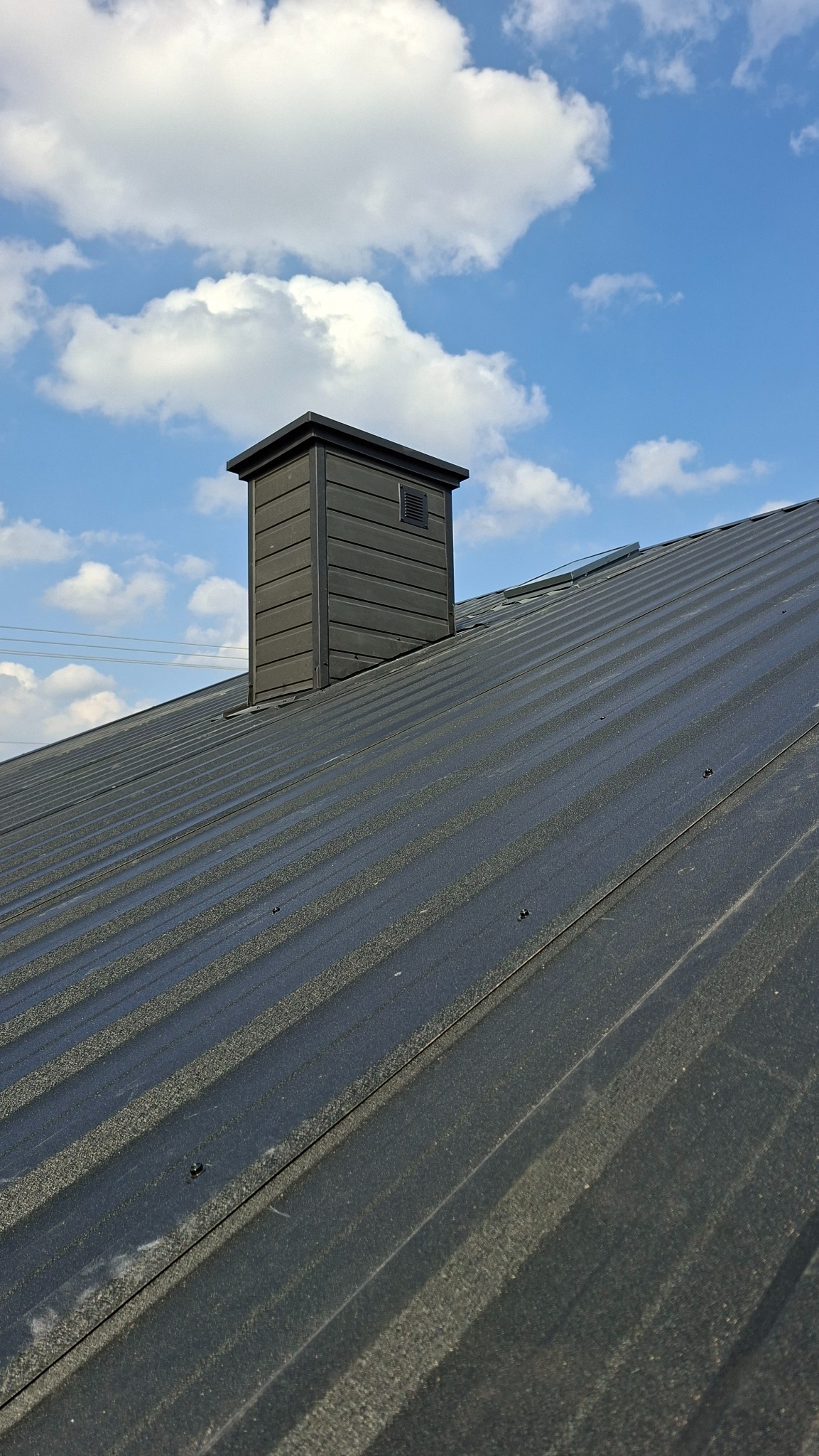 Ciemny, blaszany dach z kominem wentylacyjnym na tle błękitnego nieba z chmurami. Widoczne łączenia blachy i mocowania. Komin w kolorze dachu, o prostej, kubicznej formie.