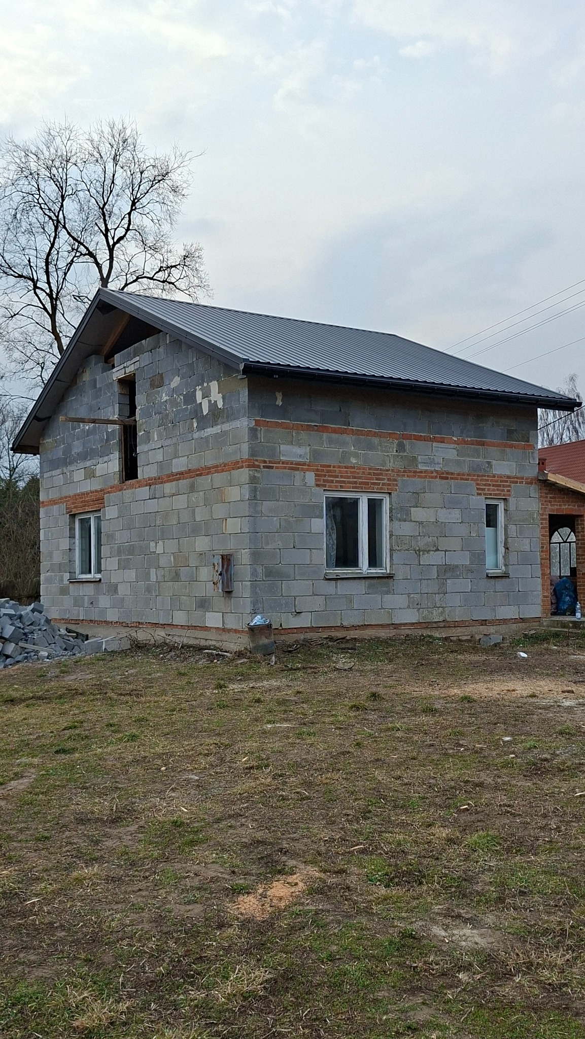 Surowy budynek z szarej bloczków i nowym, ciemnym dachem z blachy trapezowej. Widoczne okna i otwór drzwiowy. Na ziemi gruz i resztki materiałów budowlanych.