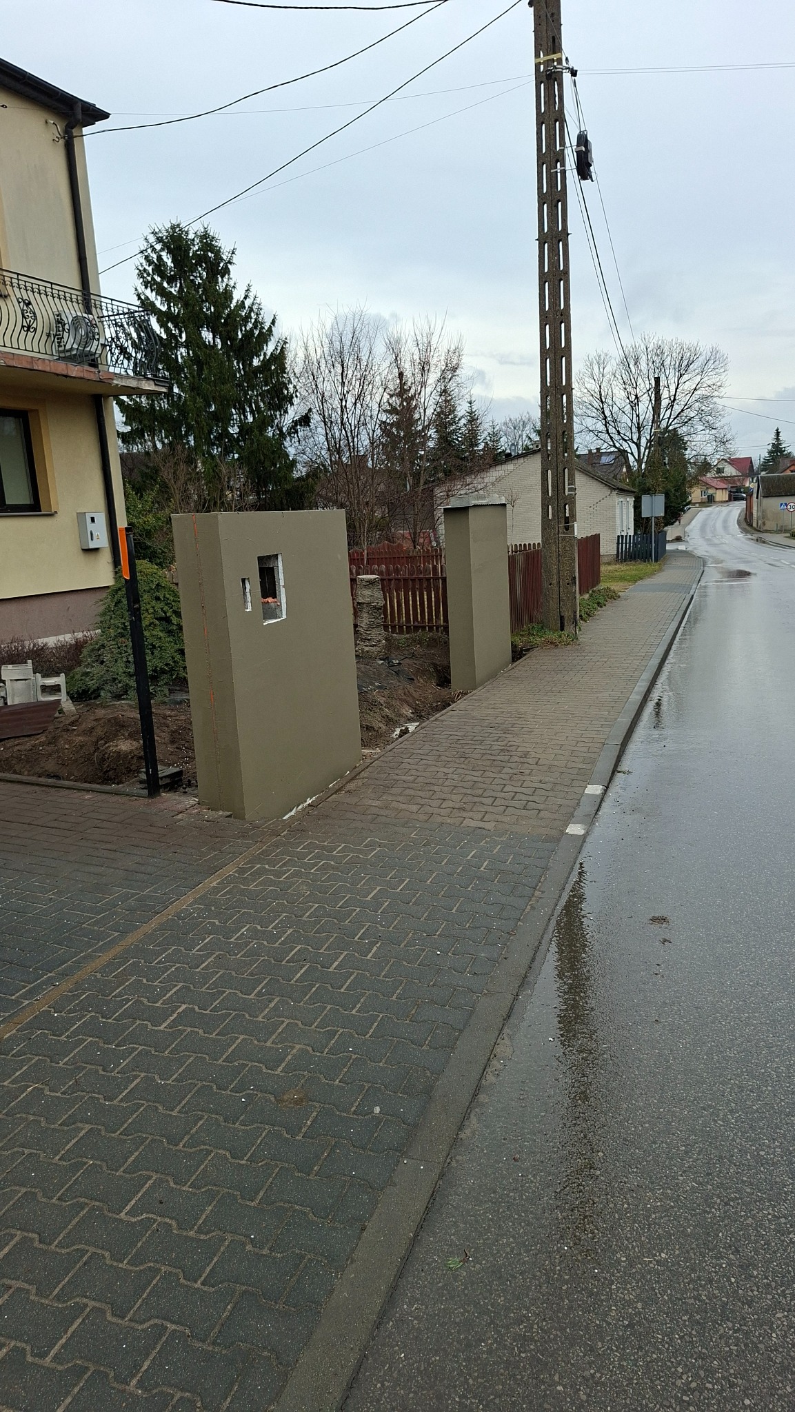 Ocieplone słupki ogrodzeniowe z małym okienkiem, widoczne z chodnika. Tło: ulica, dom i drzewa. Pochmurny dzień.