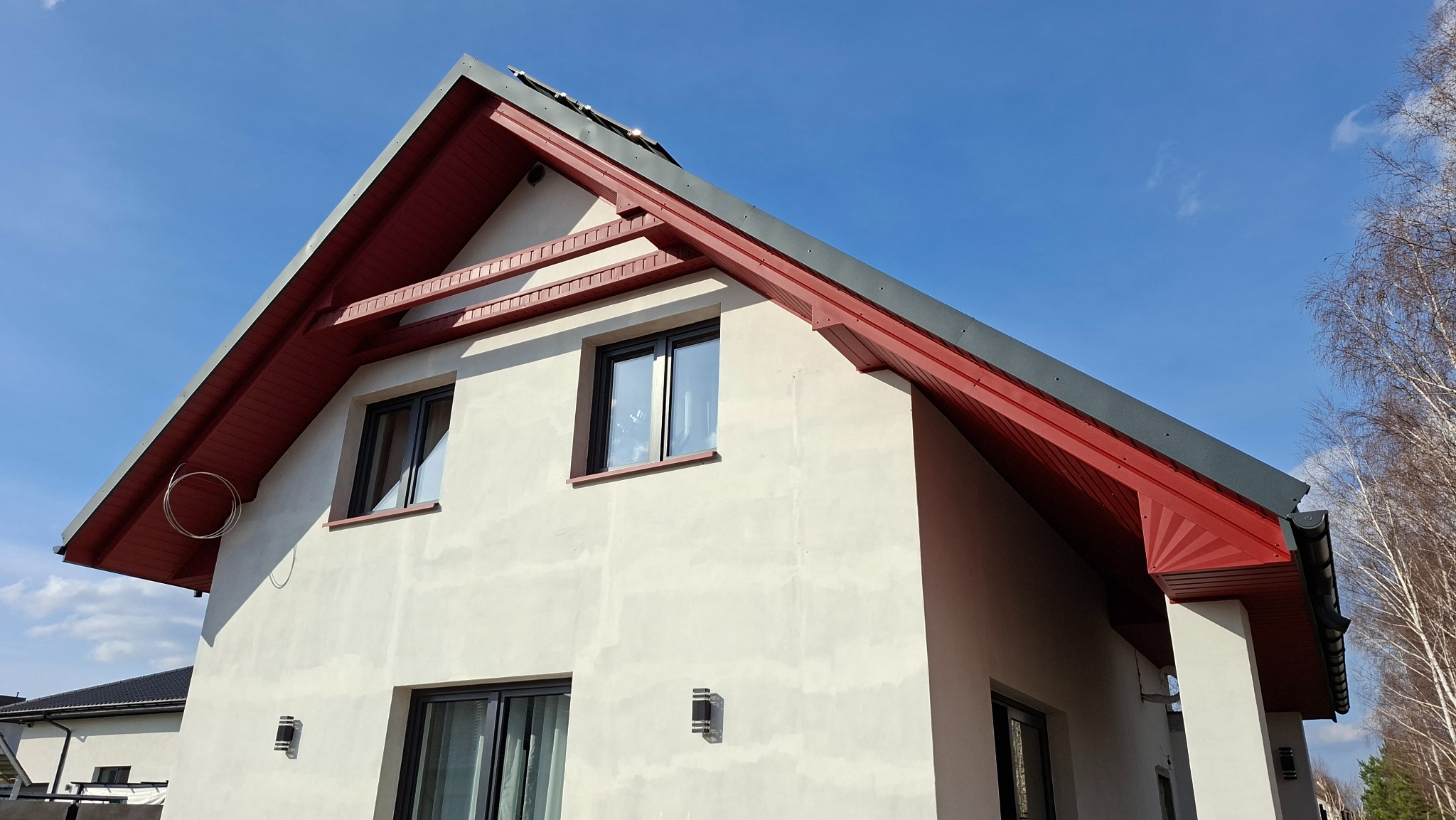 Dwukondygnacyjny dom z czerwonym dachem i szarą blachą, widziany z dołu pod kątem. Widoczne okna i elewacja w jasnym odcieniu. Błękitne niebo w tle.