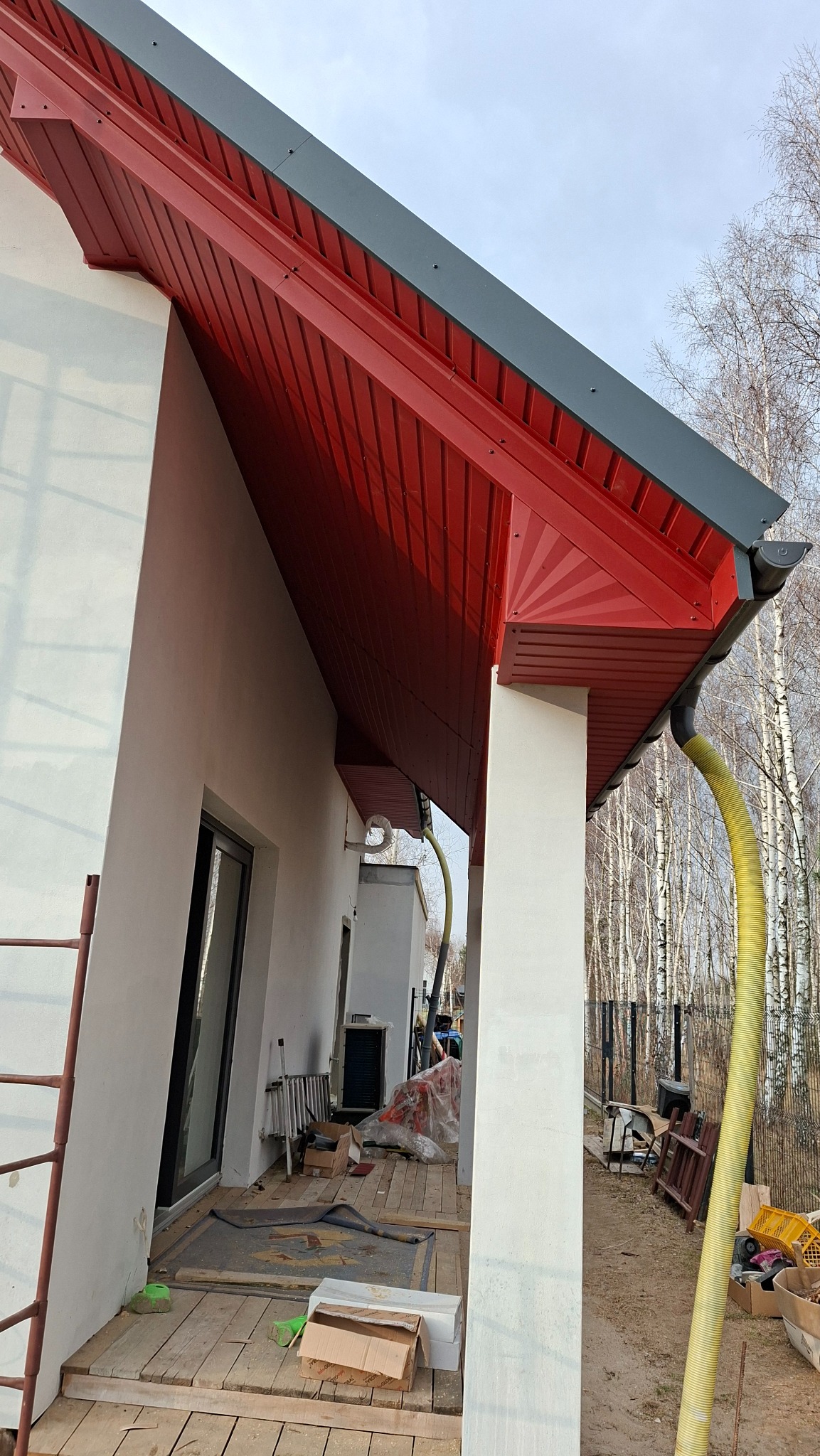 Czerwona podbitka dachu z szarą rynną na tle białej ściany budynku i brzozowego lasu. Widoczny fragment rusztowania i żółty wąż wentylacyjny.