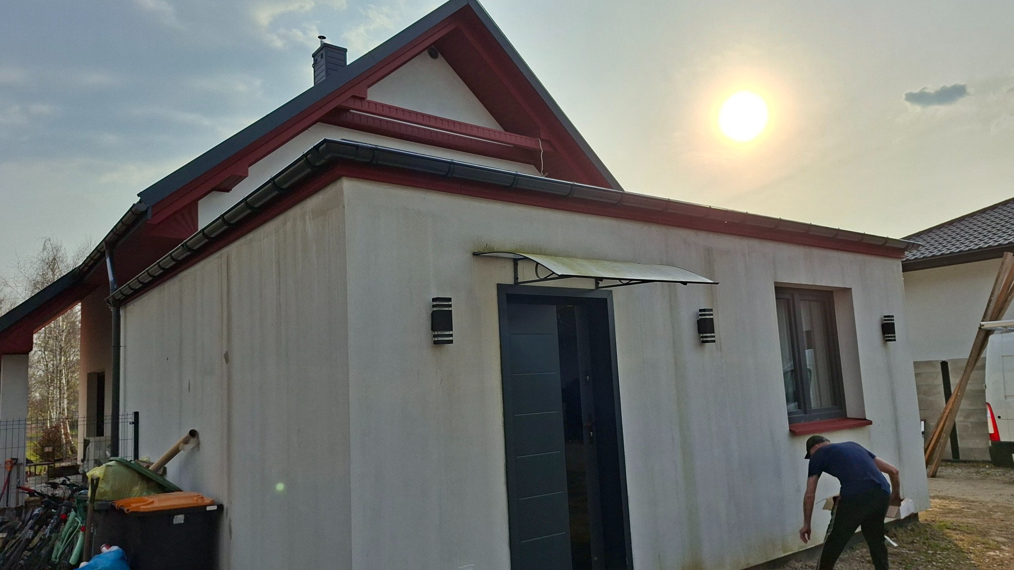 Wykończony budynek z ocieploną elewacją, widoczne rynny, dach z czerwoną podbitką i nowoczesne oświetlenie zewnętrzne. Osoba przy budynku.