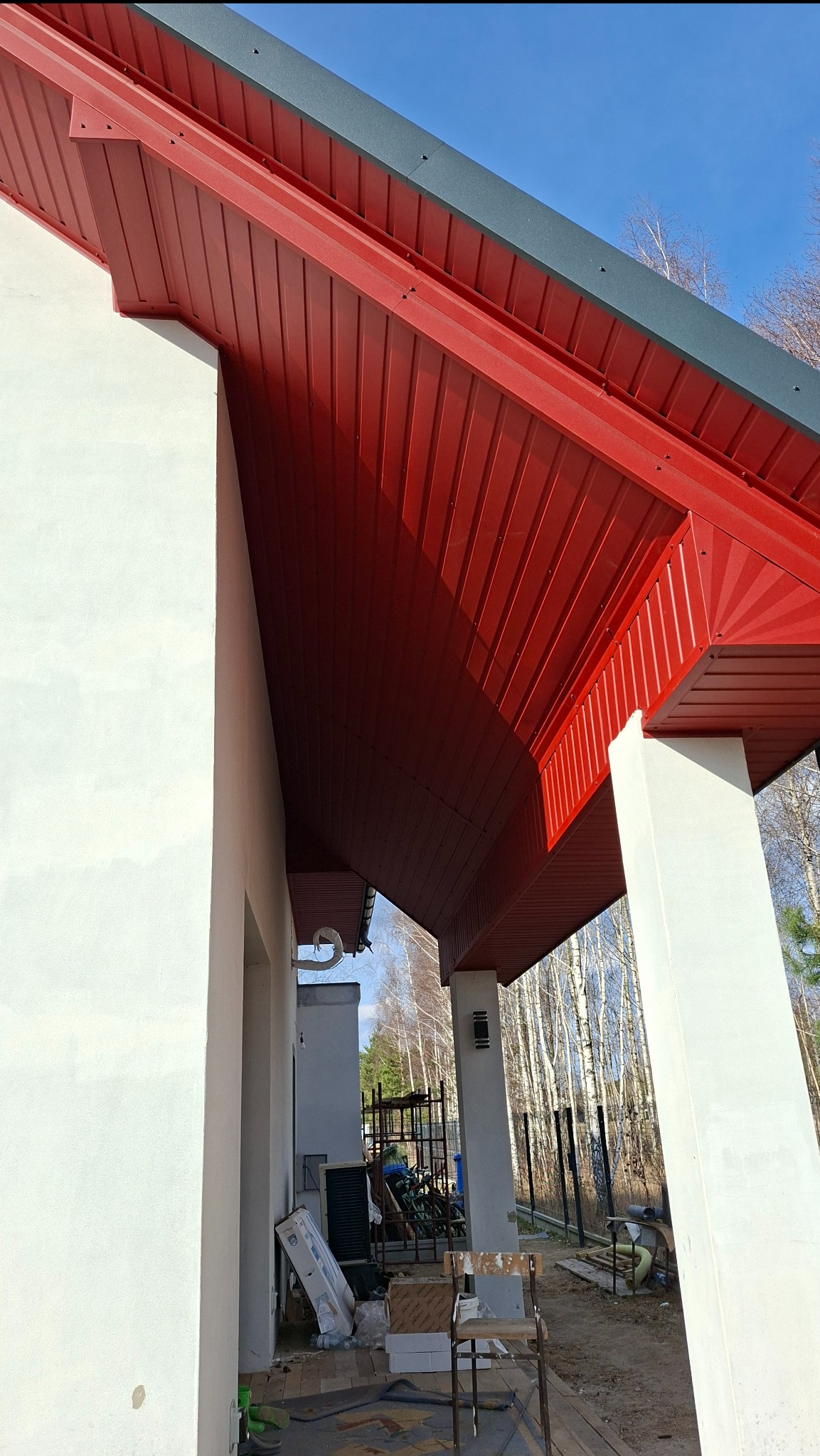 Czerwona podbitka dachowa z widocznymi elementami konstrukcyjnymi i fragmentem elewacji budynku w jasnym kolorze, na tle błękitnego nieba i drzew.