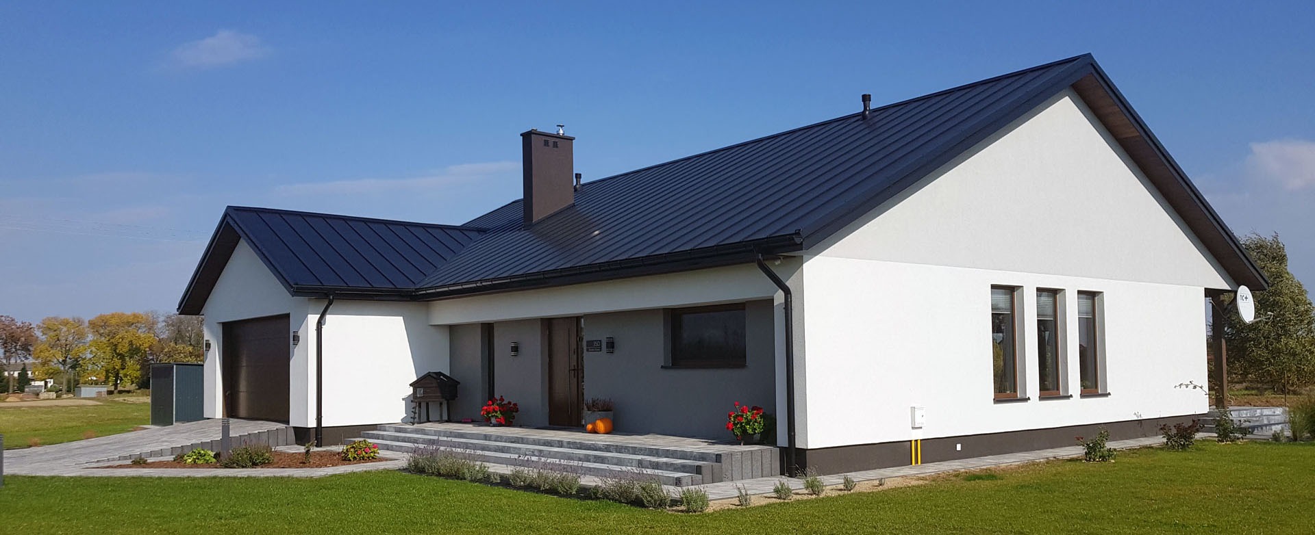 Nowoczesny dom jednorodzinny z ciemnym dachem, białymi ścianami i zadbanym trawnikiem, widok z zewnątrz w słoneczny dzień.