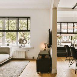 Time2Home Management - zarządzanie nieruchomościami na wynajem - Jasne, przestronne wnętrze mieszkania z drewnianą podłogą, szarą sofą, czarnymi żaluzjami w oknach i stołem jadalnym z czarnymi krzesłami.