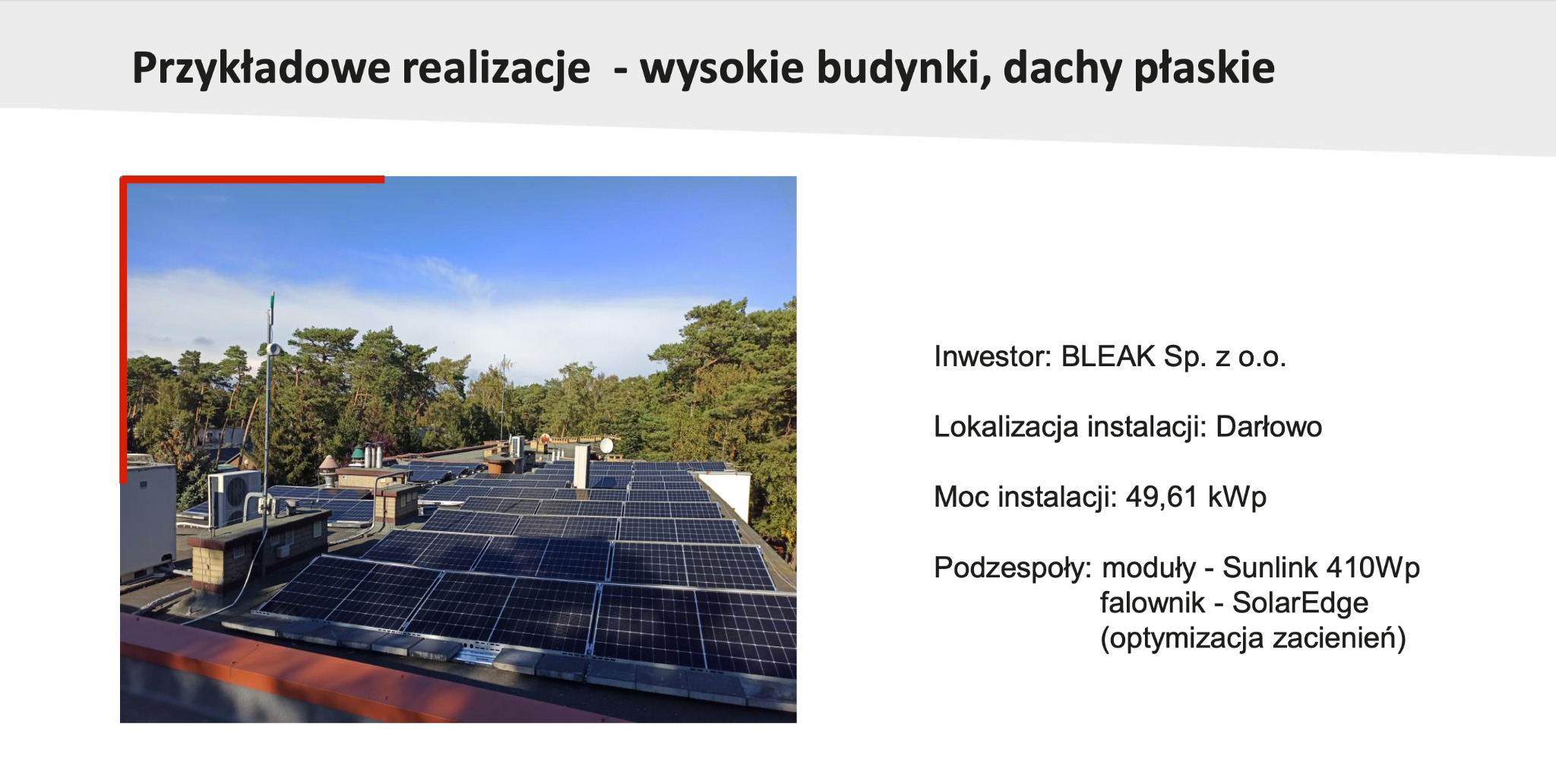 Instalacja paneli słonecznych Sunlink 410Wp z falownikiem SolarEdge na płaskim dachu wysokiego budynku w Darłowie, widok z góry na tle drzew i błękitnego nieba.