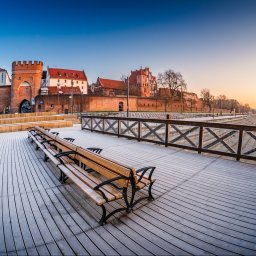 Łukasz Czajkowski - Panorama Torunia o świcie z perspektywy drewnianego molo z ławkami. W tle brama miejska, mury obronne i most na Wiśle w promieniach wschodzącego słońca.