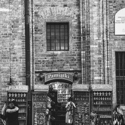 Łukasz Czajkowski - Czarno-białe zdjęcie fasady sklepu z pamiątkami w zabytkowej kamienicy. Widoczne regały z pamiątkami, okno i fragment tablicy informacyjnej. Osoba przygląda się towarom.