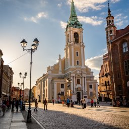 Łukasz Czajkowski - Słoneczny kadr z perspektywy ulicy na kościół z zieloną wieżą w Toruniu. Widoczni przechodnie i latarnie. Ciepłe światło podkreśla architekturę.