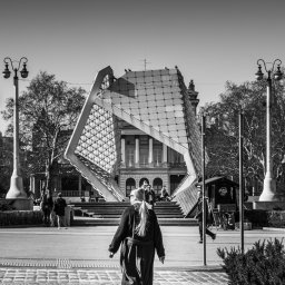 Łukasz Czajkowski - Czarno-białe zdjęcie kobiety idącej w stronę nowoczesnej fontanny na tle klasycystycznego budynku. Ujęcie z perspektywy pieszego, w słoneczny dzień.