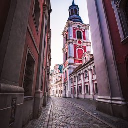 Łukasz Czajkowski - Ujęcie z dołu na barokowy kościół z czerwoną fasadą i wieżą, widziany z brukowanej uliczki w Poznaniu, Polska. Architektura miejska w ciepłych barwach.