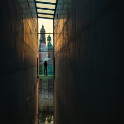 Łukasz Czajkowski - Wąski korytarz z przeszklonym dachem i podłogą, przez które widać sylwetkę osoby i fragmenty architektury miejskiej w ciepłym świetle zachodzącego słońca.