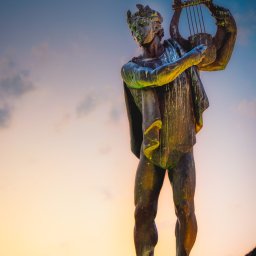 Łukasz Czajkowski - Statua mężczyzny z lirą na tle nieba o zachodzie słońca. Niskie ujęcie podkreśla monumentalność rzeźby i ciepłe barwy zachodzącego słońca.