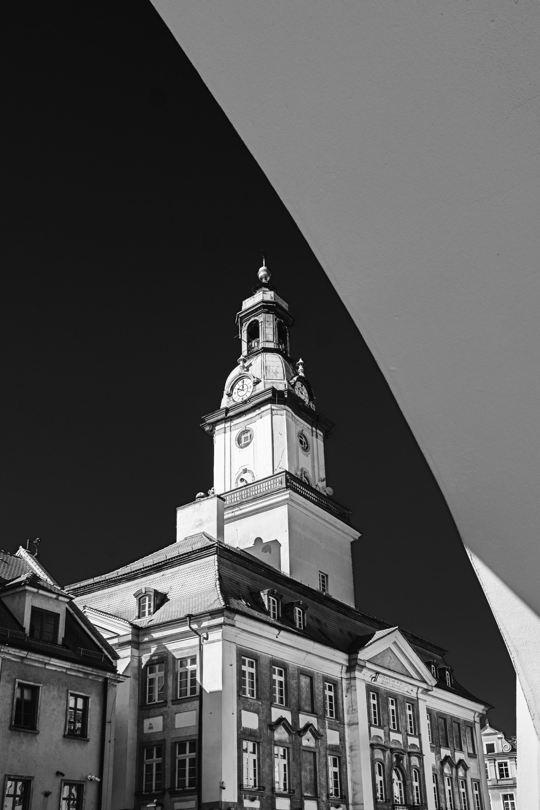 Czarno-białe zdjęcie wieży zegarowej z budynkiem w tle, widzianej z dołu, z fragmentem łuku w prawym górnym rogu kadru, tworzącym geometryczną kompozycję.