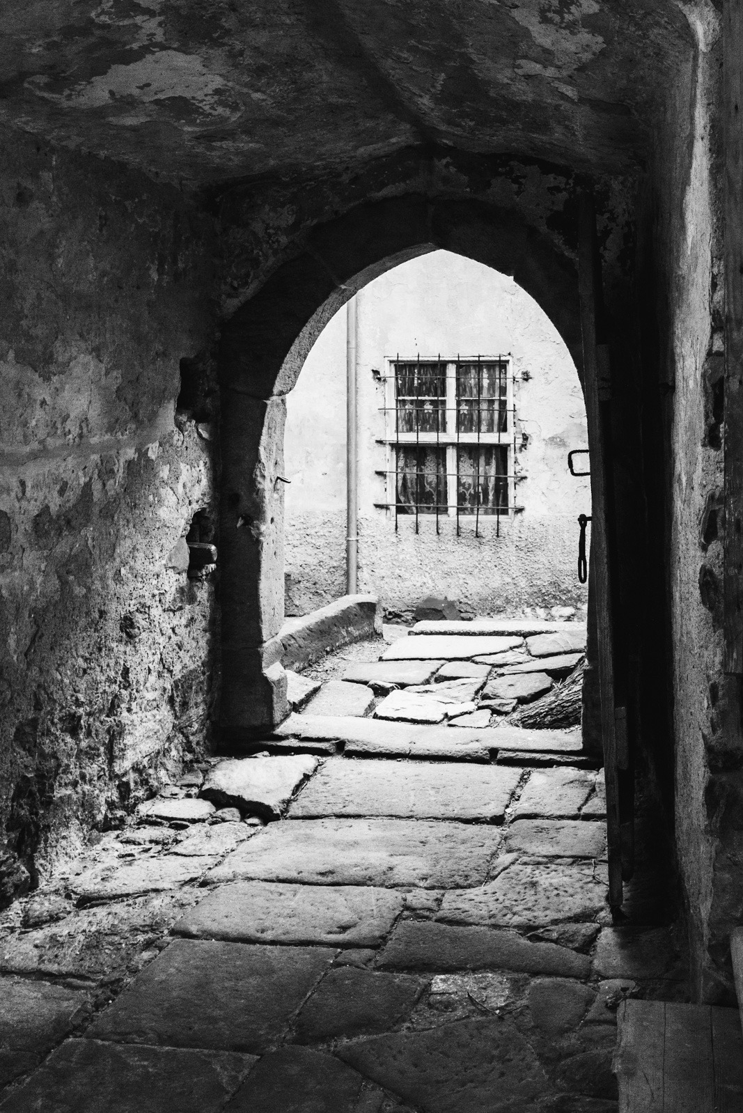 Czarno-białe zdjęcie kamiennej uliczki widzianej przez łukowate przejście. W tle okno z kratami i fragment budynku. Surowy, historyczny klimat.
