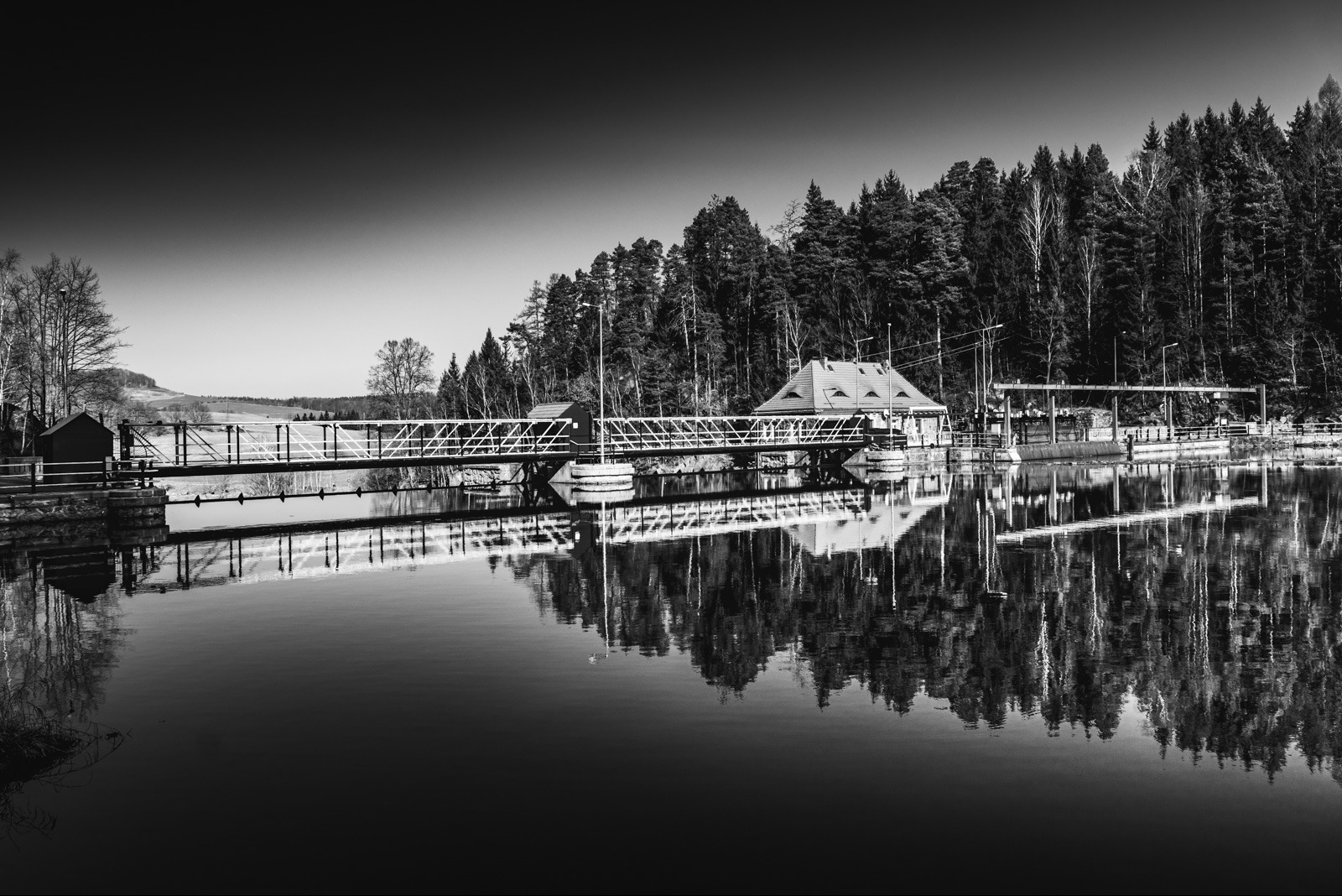 Czarno-białe zdjęcie tamy z budynkiem i mostem, odbijającej się w spokojnej wodzie. W tle las iglasty pod pochmurnym niebem.