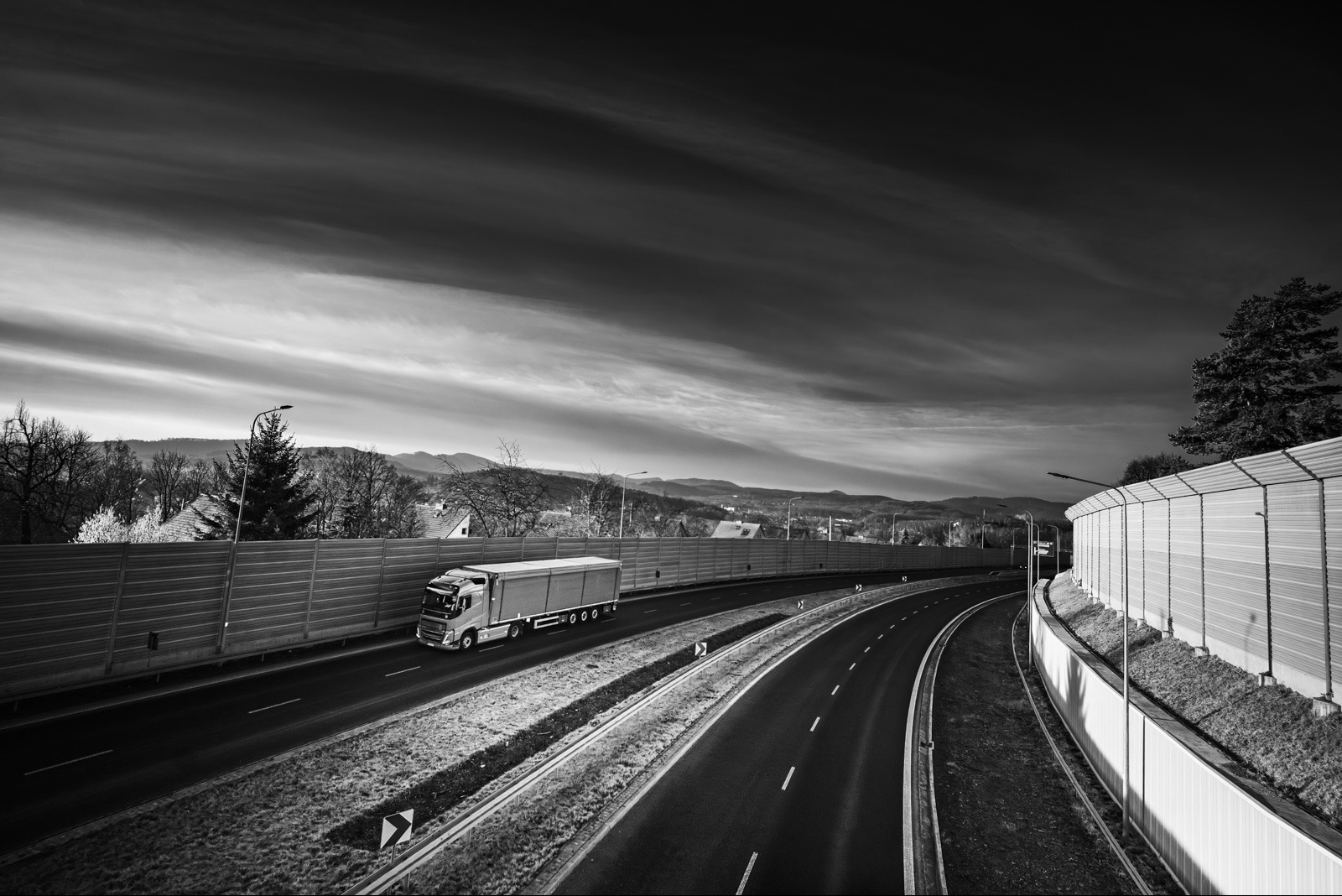 Czarno-białe zdjęcie ciężarówki na autostradzie, z widokiem na góry w tle i dynamicznym niebem. Ujęcie z góry, podkreślające perspektywę drogi.