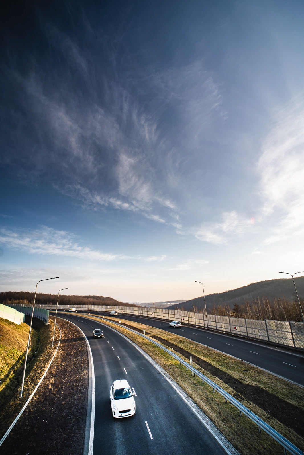 Szeroki kadr autostrady z białym samochodem w ruchu, błękitne niebo z chmurami, bariery dźwiękochłonne i zalesione wzgórza w tle, wczesny poranek.